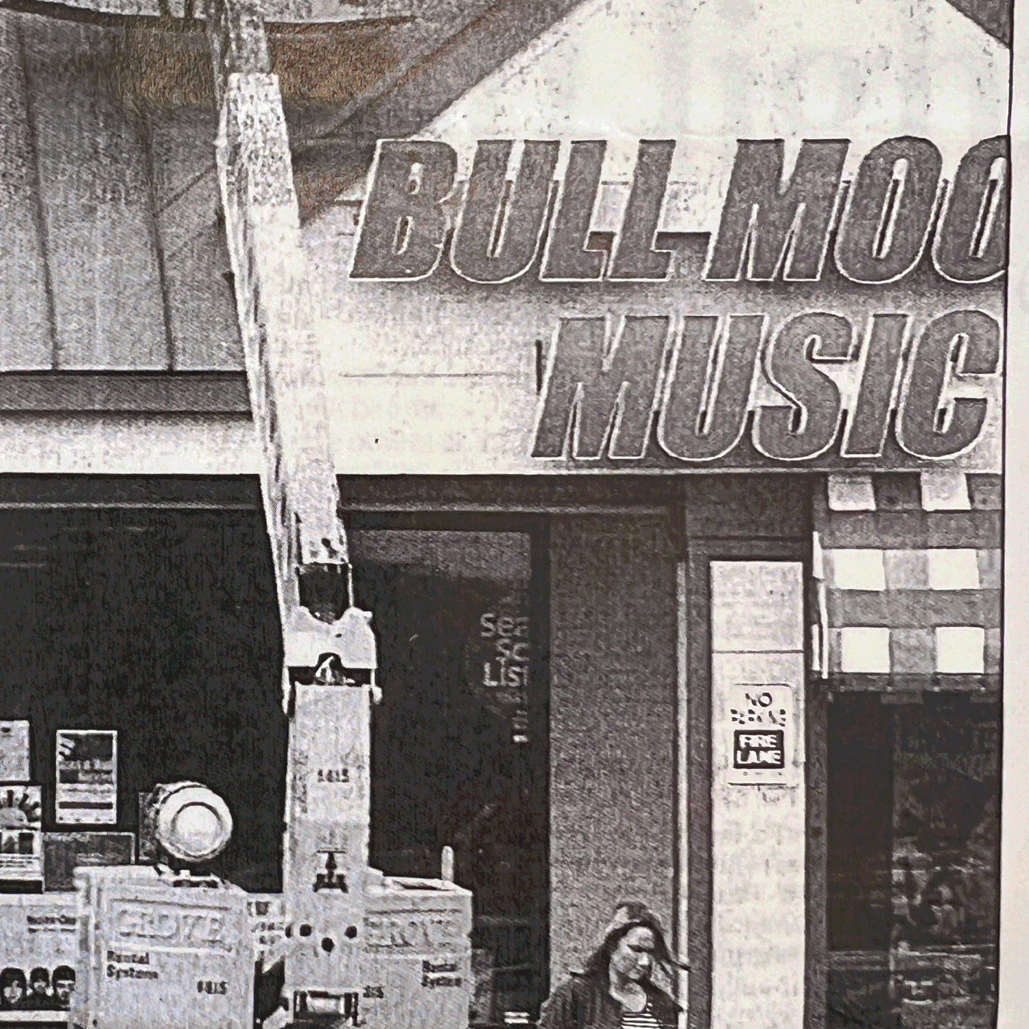 Black and white photo of a music store with a large sign that reads 'BULL MOOSE MUSIC', a person with long hair and glasses walking past, and various posters and items displayed outside the store.