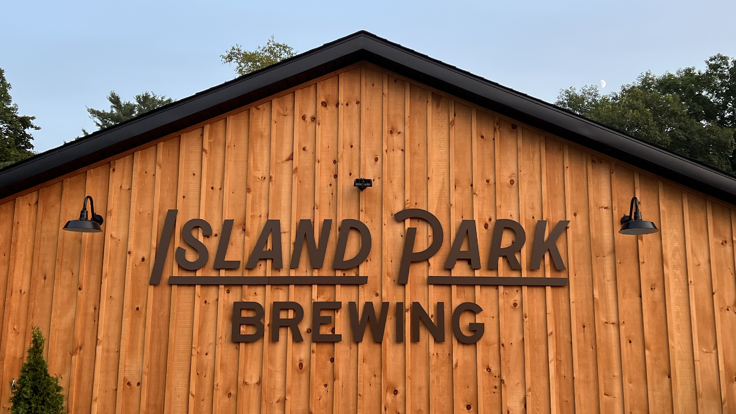 Wooden building with a sign reading "Island Park Brewing" and two black wall-mounted lights, trees in the background, clear sky with the moon visible.