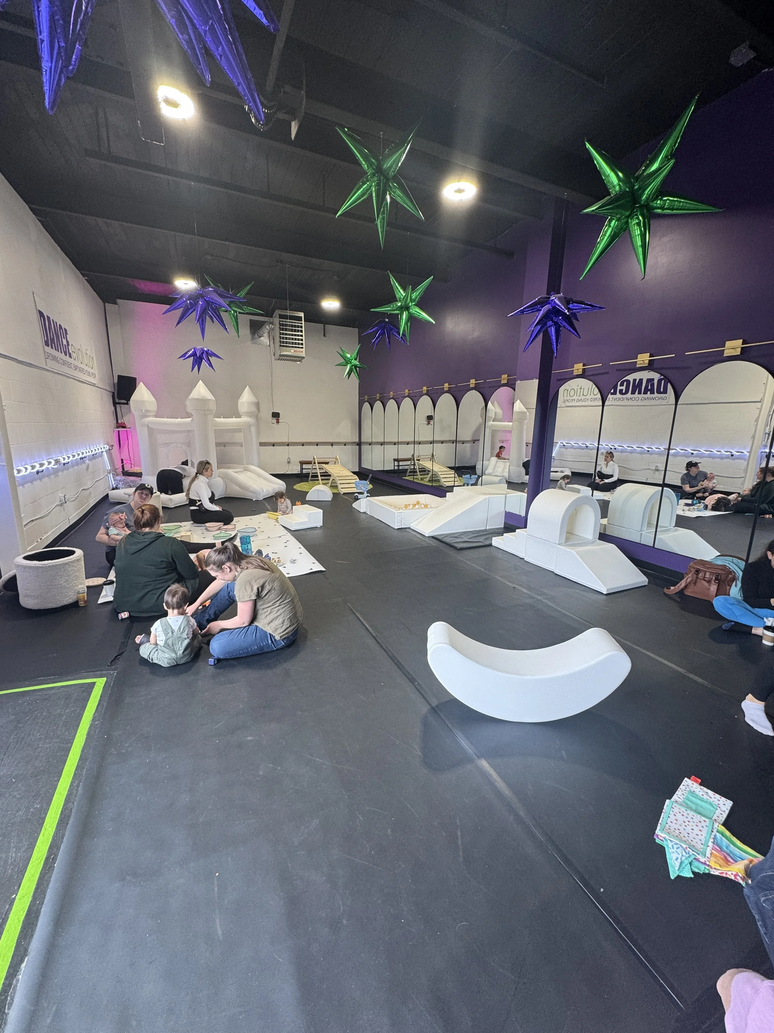 Indoor play area with black flooring, white and purple decor, large star-shaped hanging lights in green and purple, seating for adults, and children playing on mats with toys.