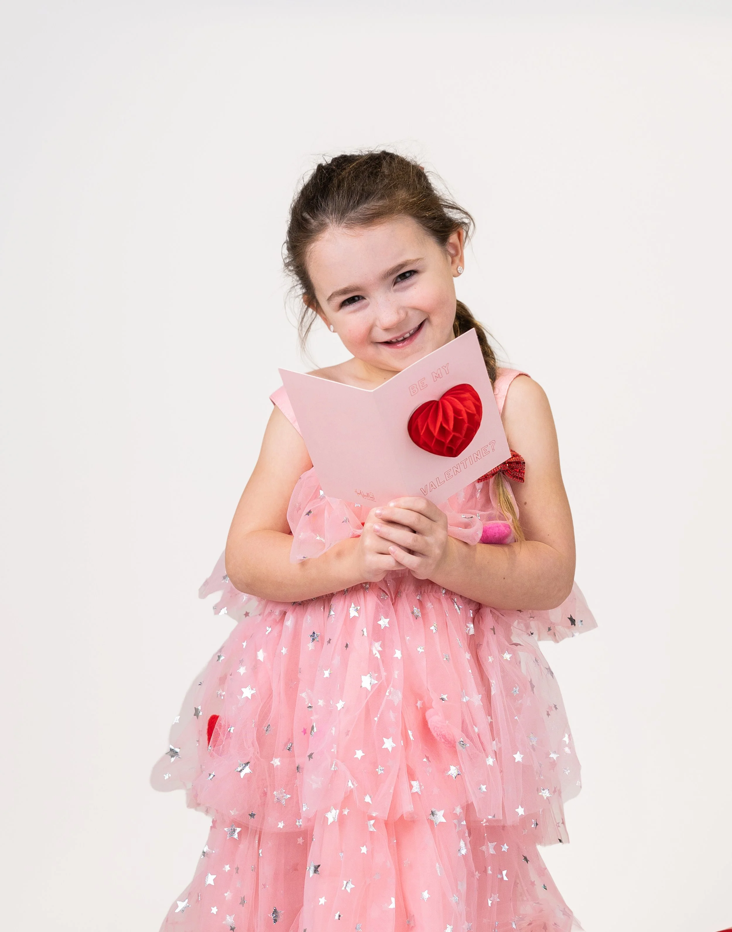 A young girl wearing a pink dress with silver stars, holding a Valentine's Day card with a red heart, smiling against a white background.