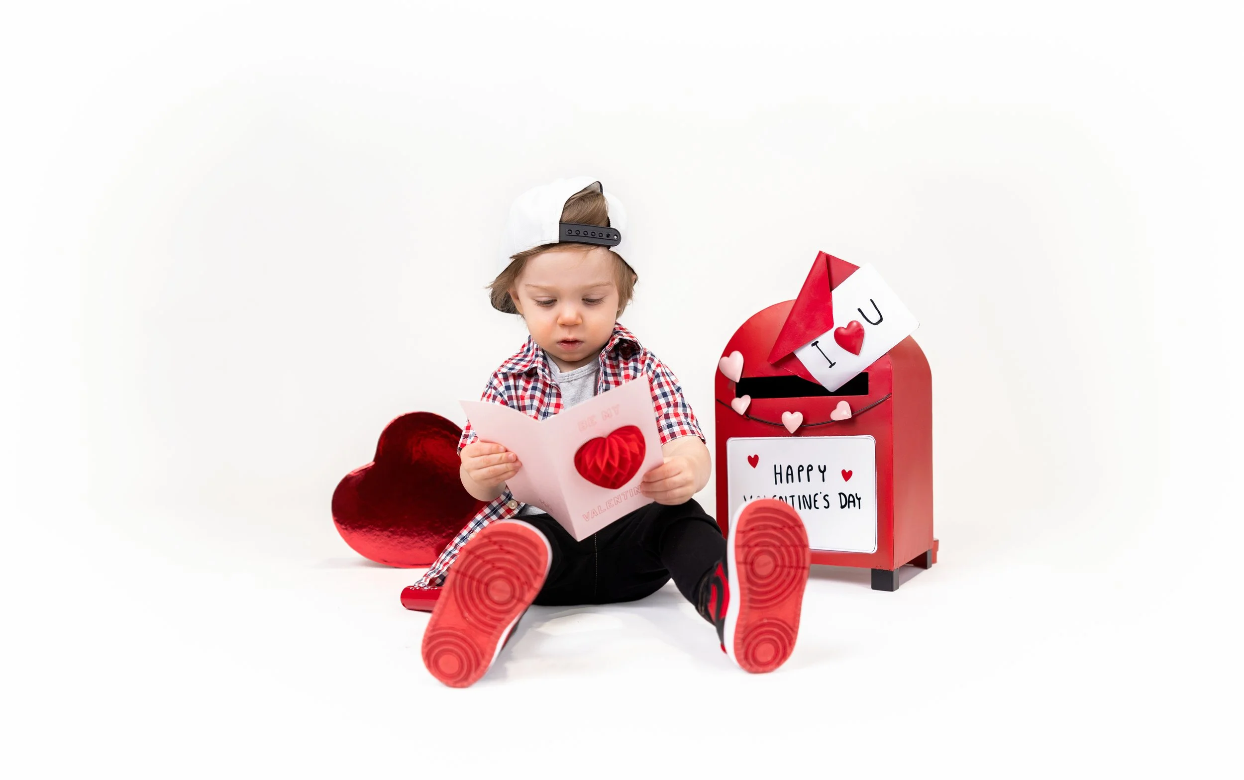 A young boy sitting on the floor, reading a Valentine's Day card, surrounded by Valentine's Day decorations including a red mailbox, heart-shaped pillows, and a sign that says 'Happy Valentine's Day.'