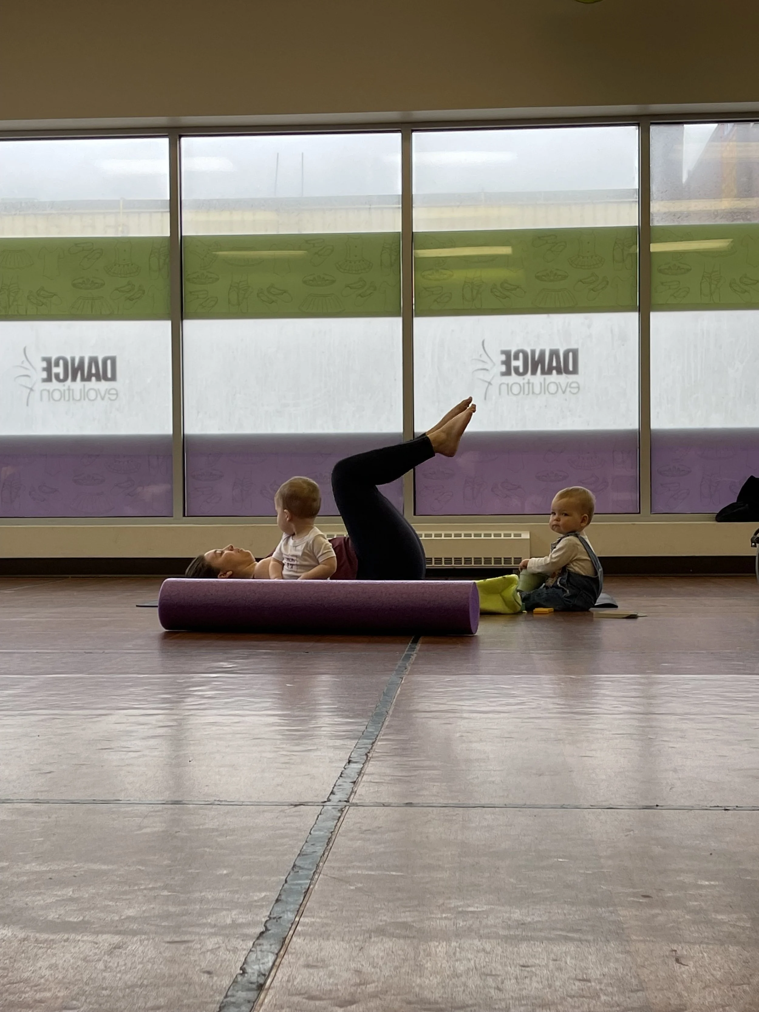 A woman lying on her back on a purple yoga mat, using her legs to hold a small child on her stomach while another small child sits nearby on the floor, inside a fitness or yoga studio with large windows.