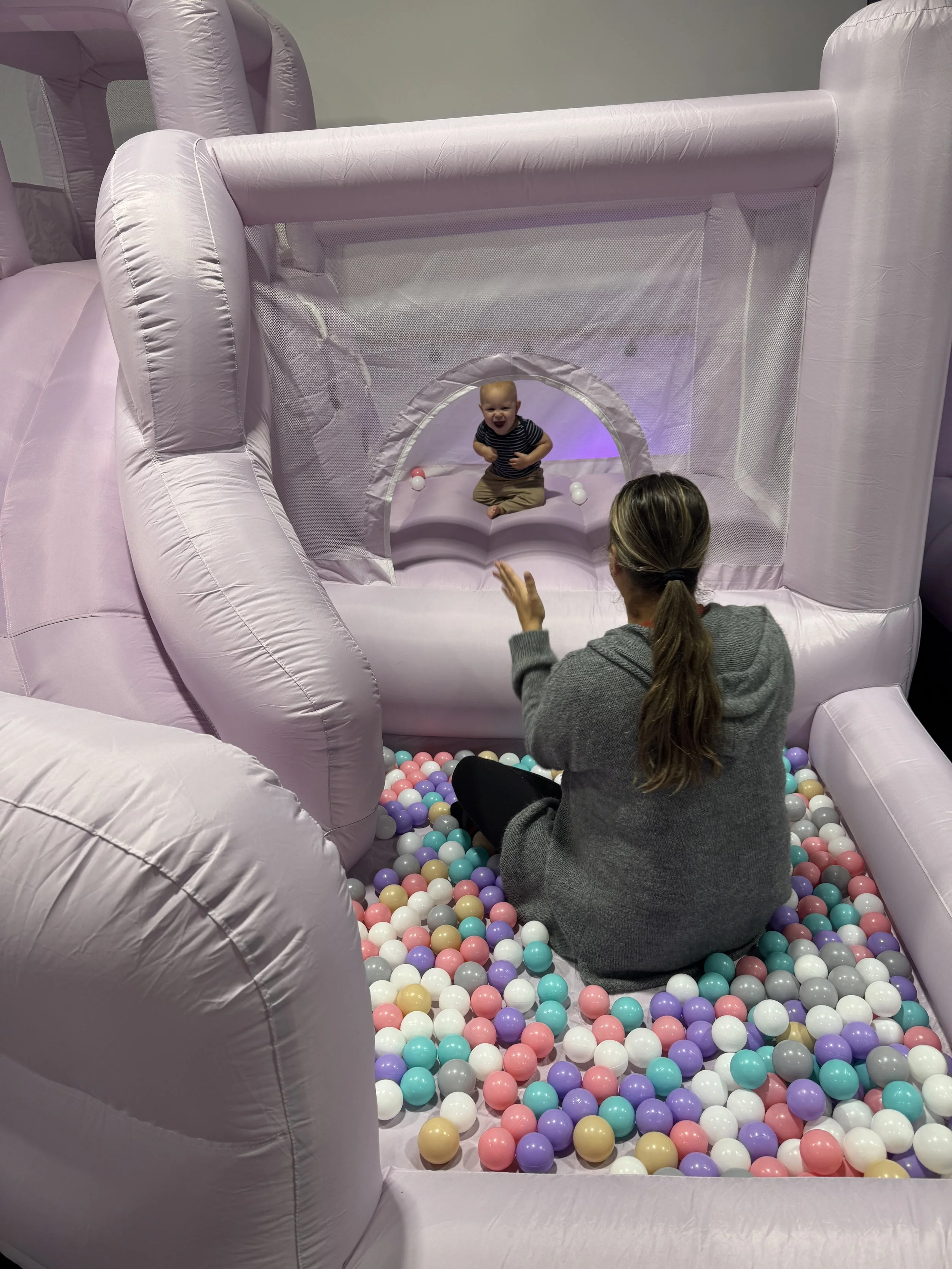 A young child sitting in a ball pit, laughing and playing with an adult, inside an inflatable pink play area with a tunnel and colorful balls.