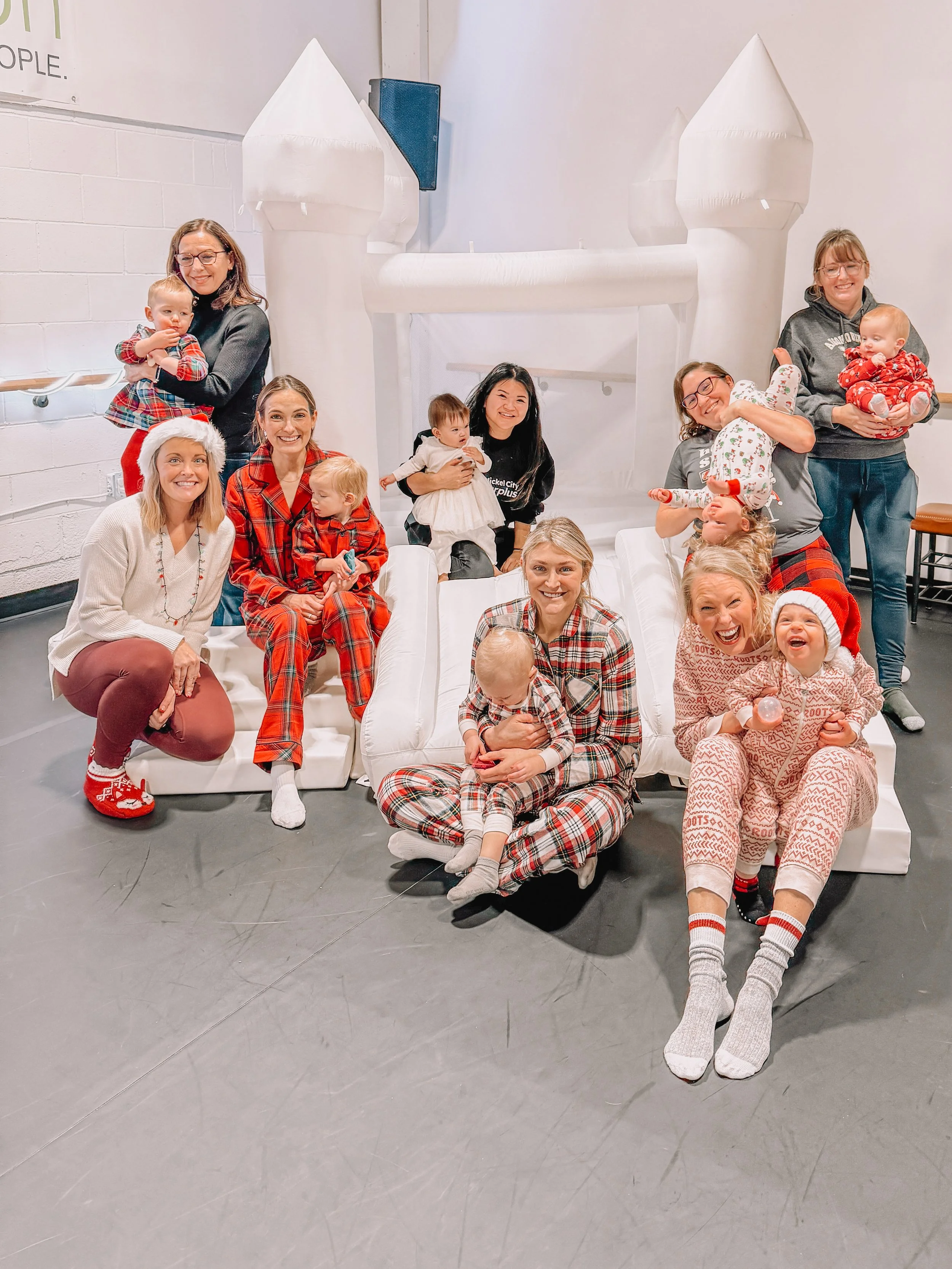 Group of women and children celebrating Christmas with festive pajamas, Santa hats, and laughing together in front of a white inflatable castle.