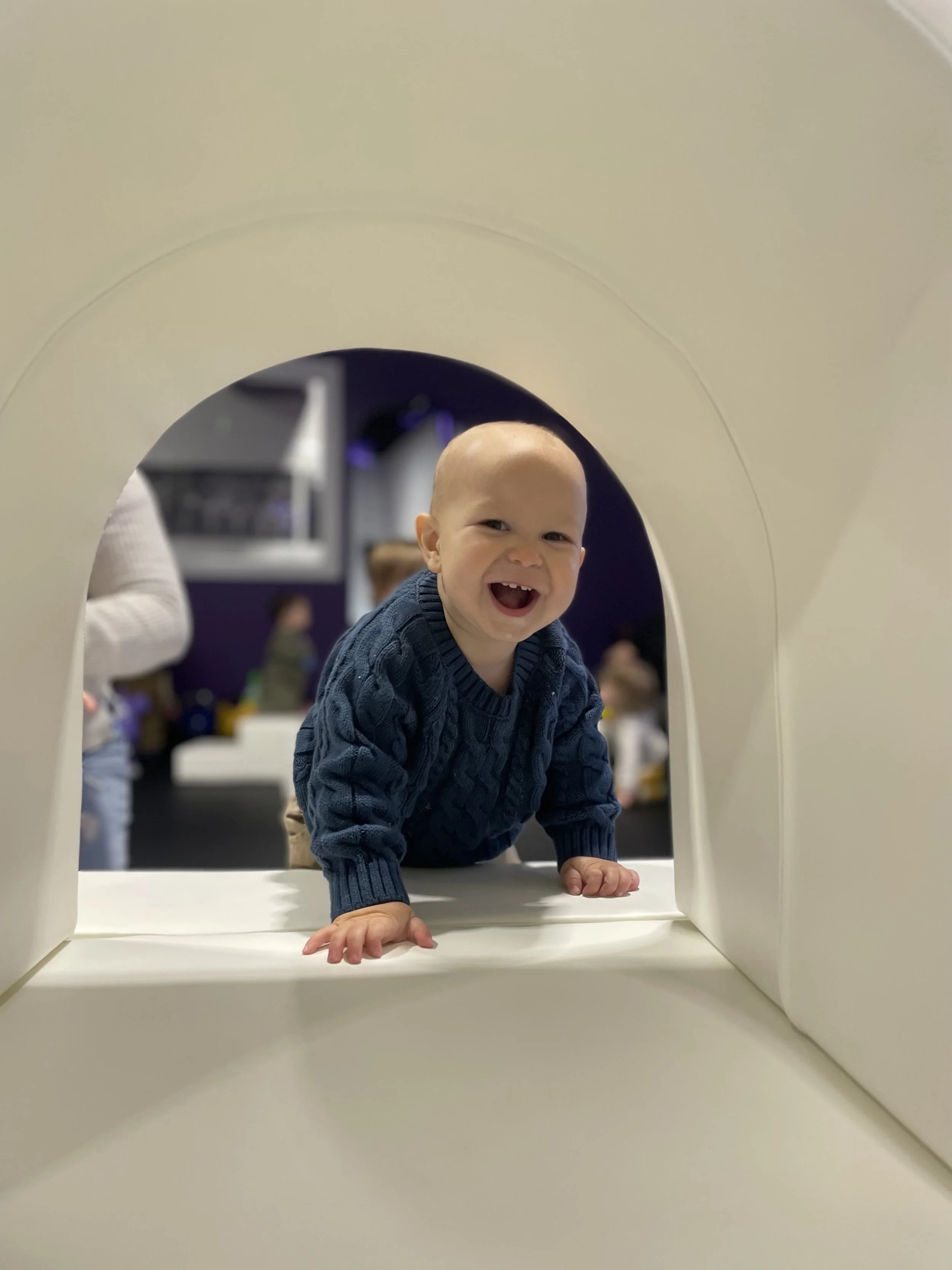 A young boy with a big smile crawling through a white play tunnel in an indoor play area.