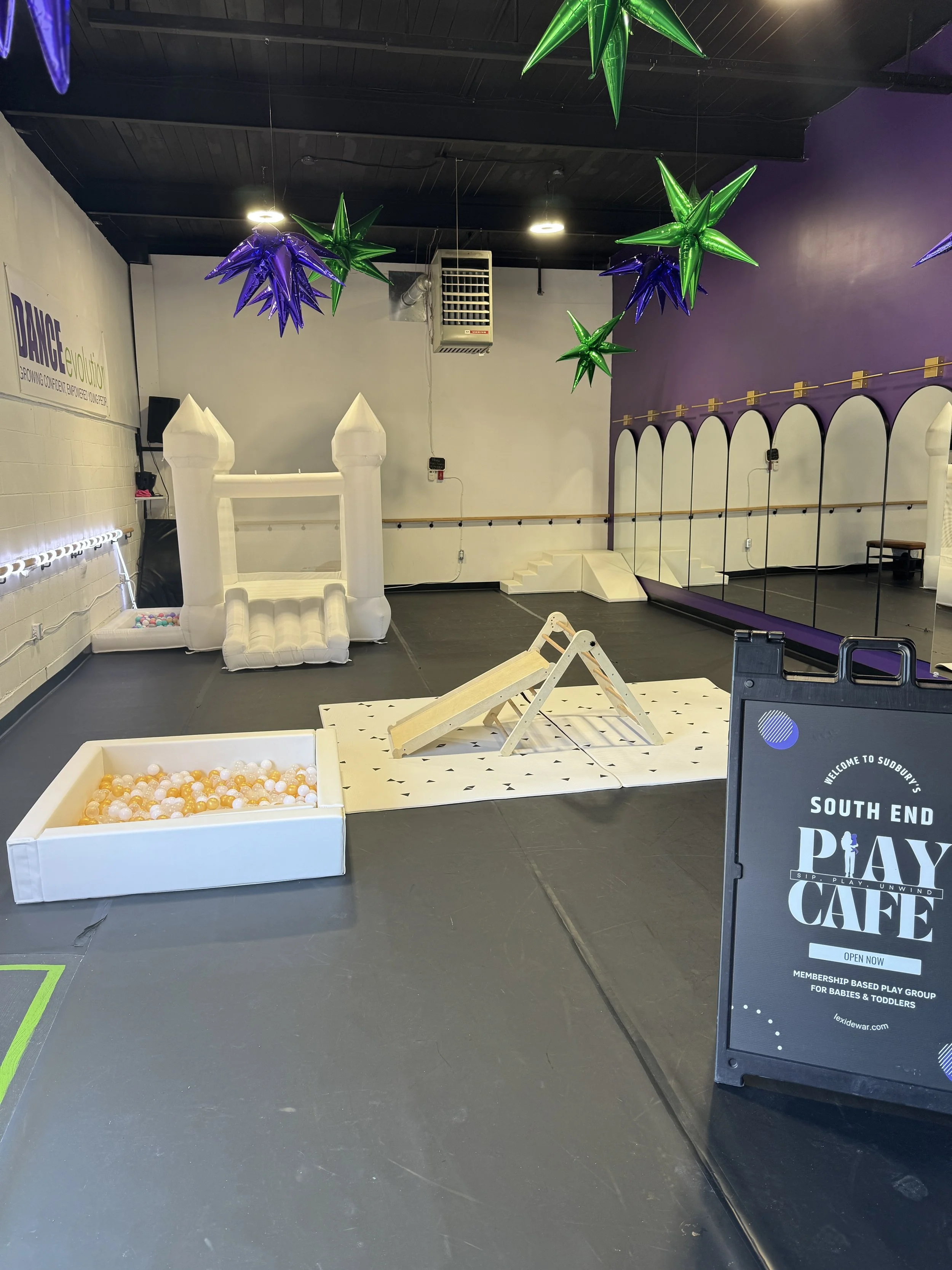 Indoor play area with a white inflatable castle, a sandbox with plastic balls, a small slide, colorful hanging star-shaped decorations, and a sign for South End Play Cafe for babies and toddlers.