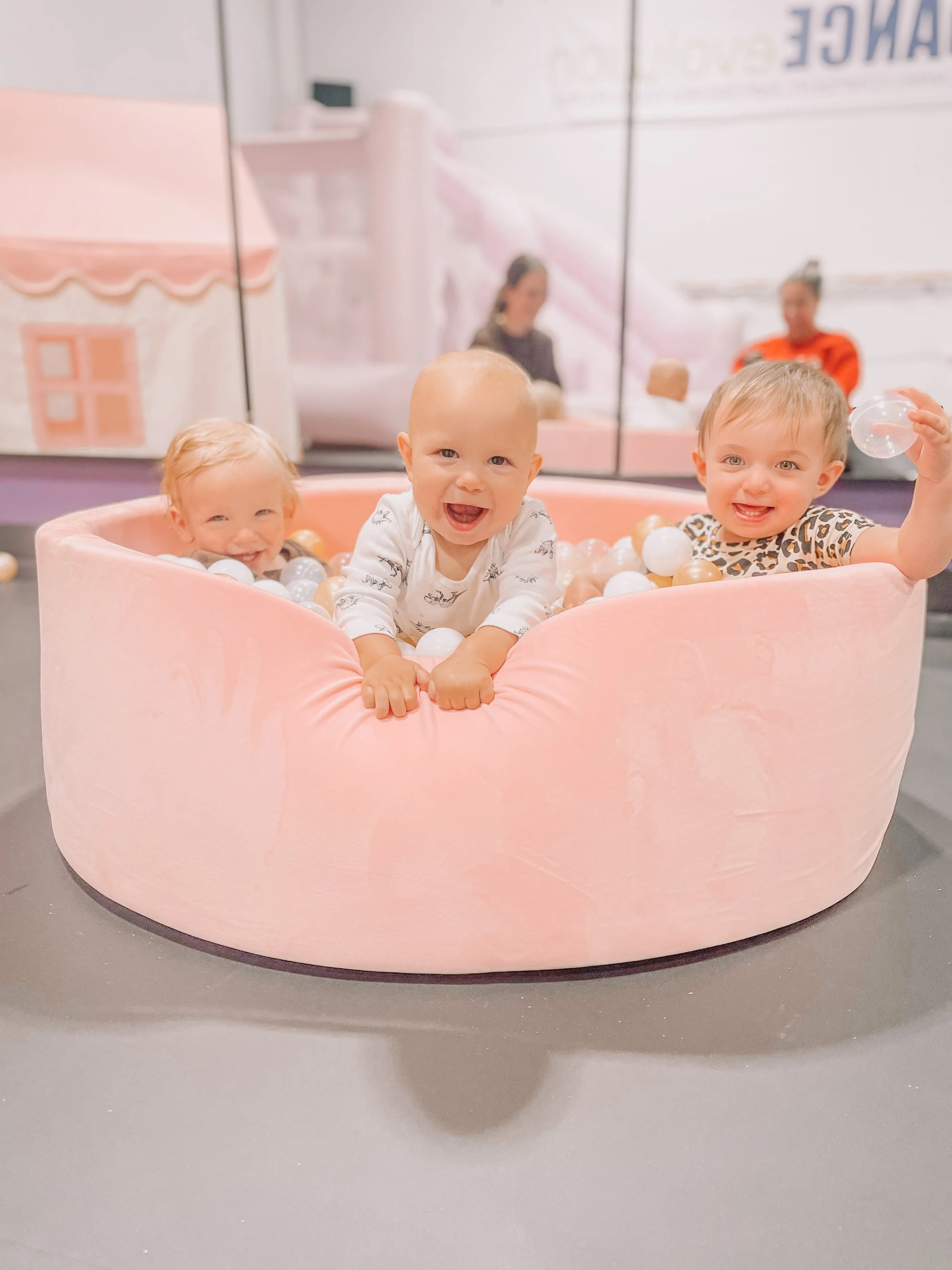 Three babies smiling and playing in a pink ball pit, with a mirror and adults in the background.