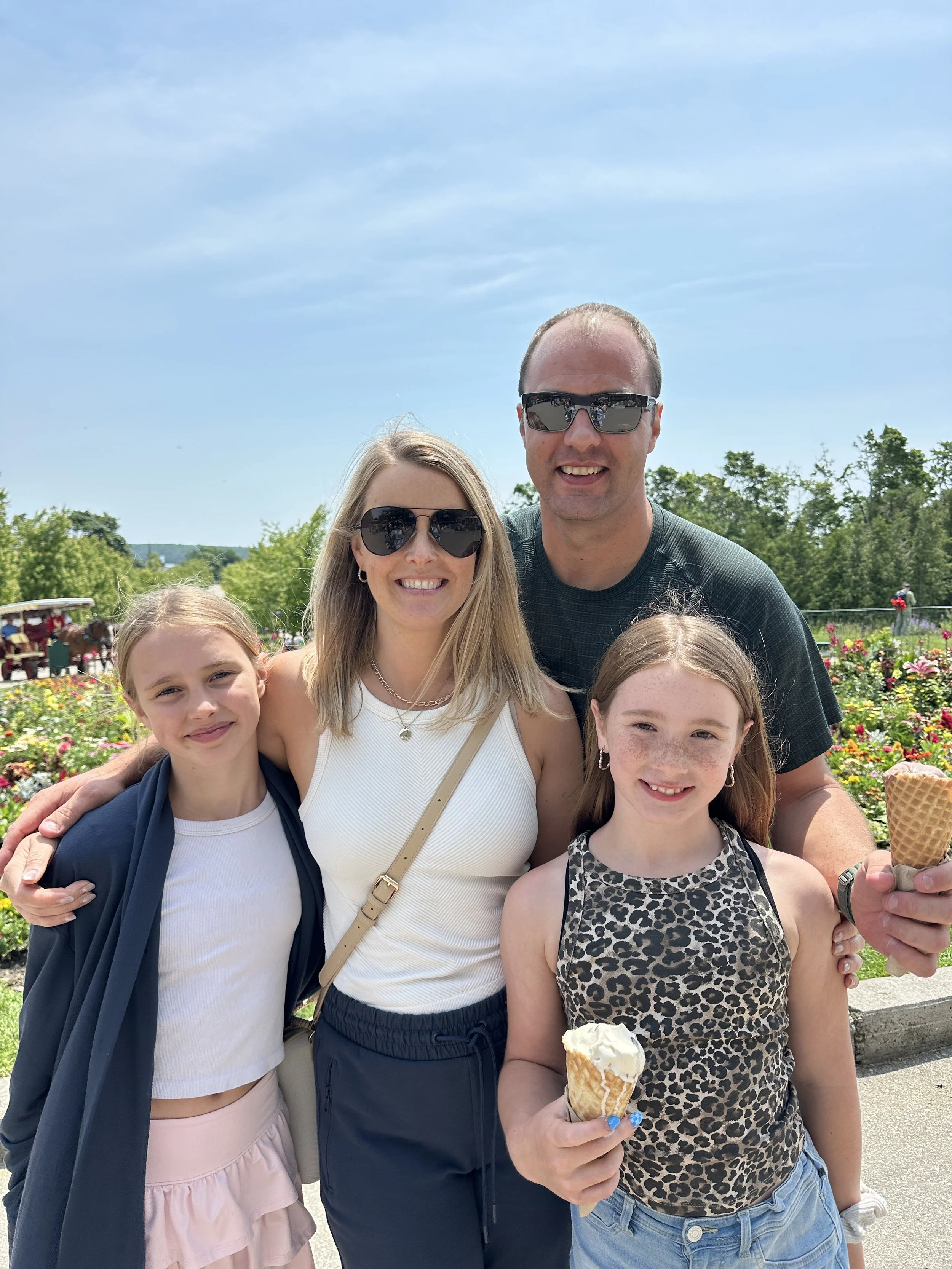 A family of four with two young girls, a woman, and a man, smiling outdoors with a garden of colorful flowers behind them, each holding an ice cream cone, on a sunny day.