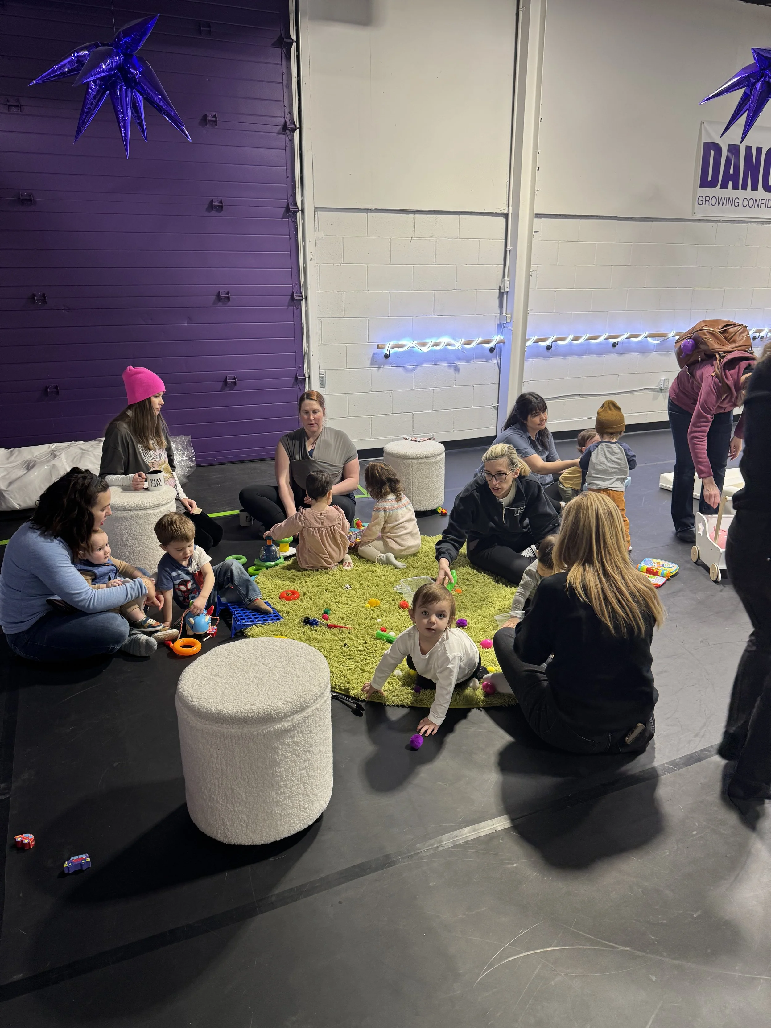 Children and adults sitting on a green rug in a dance studio, surrounded by toys, with purple and white walls, and a purple door in the background.