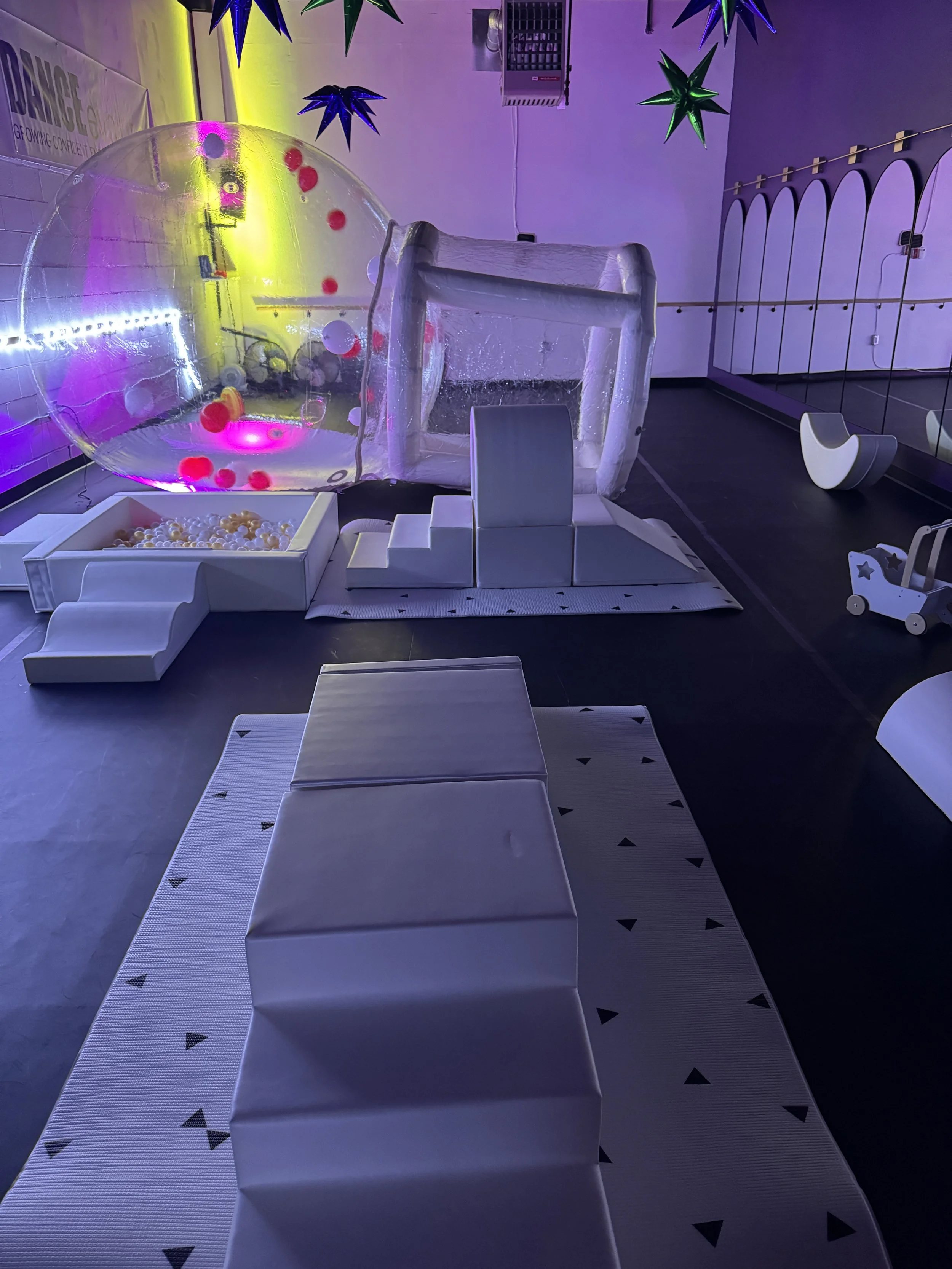 Indoor play area with inflatable ball pit, inflatable bounce house, and soft seating. Decorated with star-shaped hanging mobiles and colorful lighting.