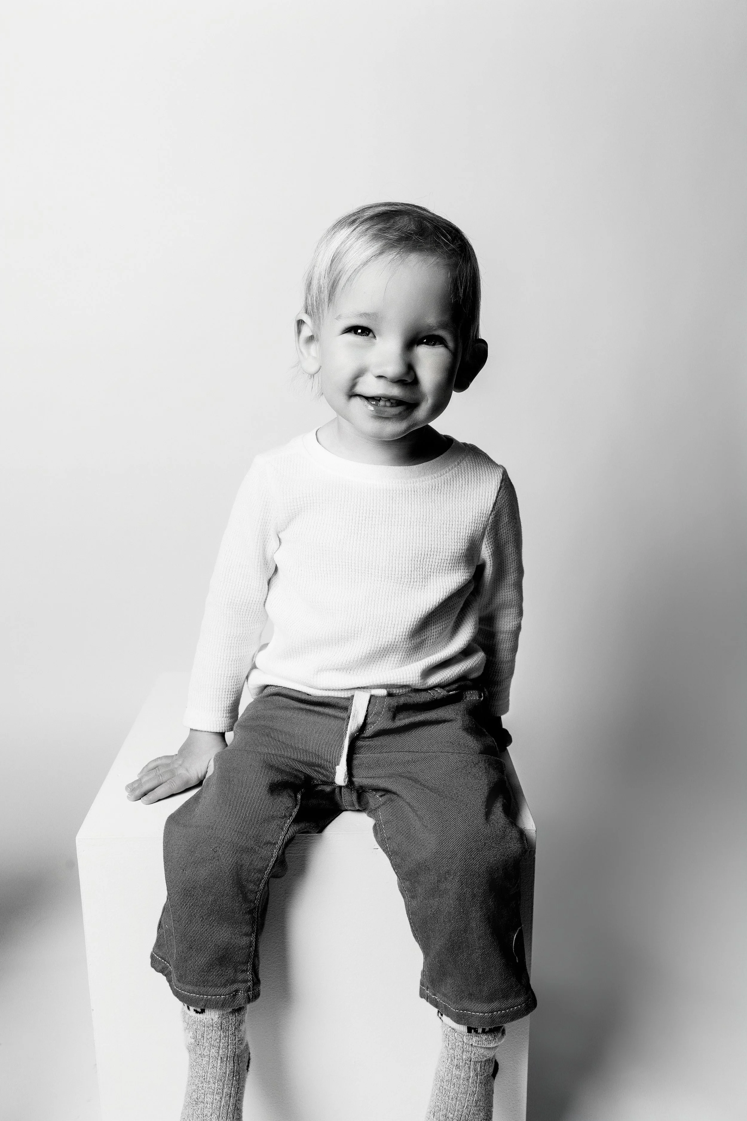 A smiling young boy with light hair, wearing a white long-sleeve shirt, dark pants, and socks, sitting on a white block against a plain background in black and white.