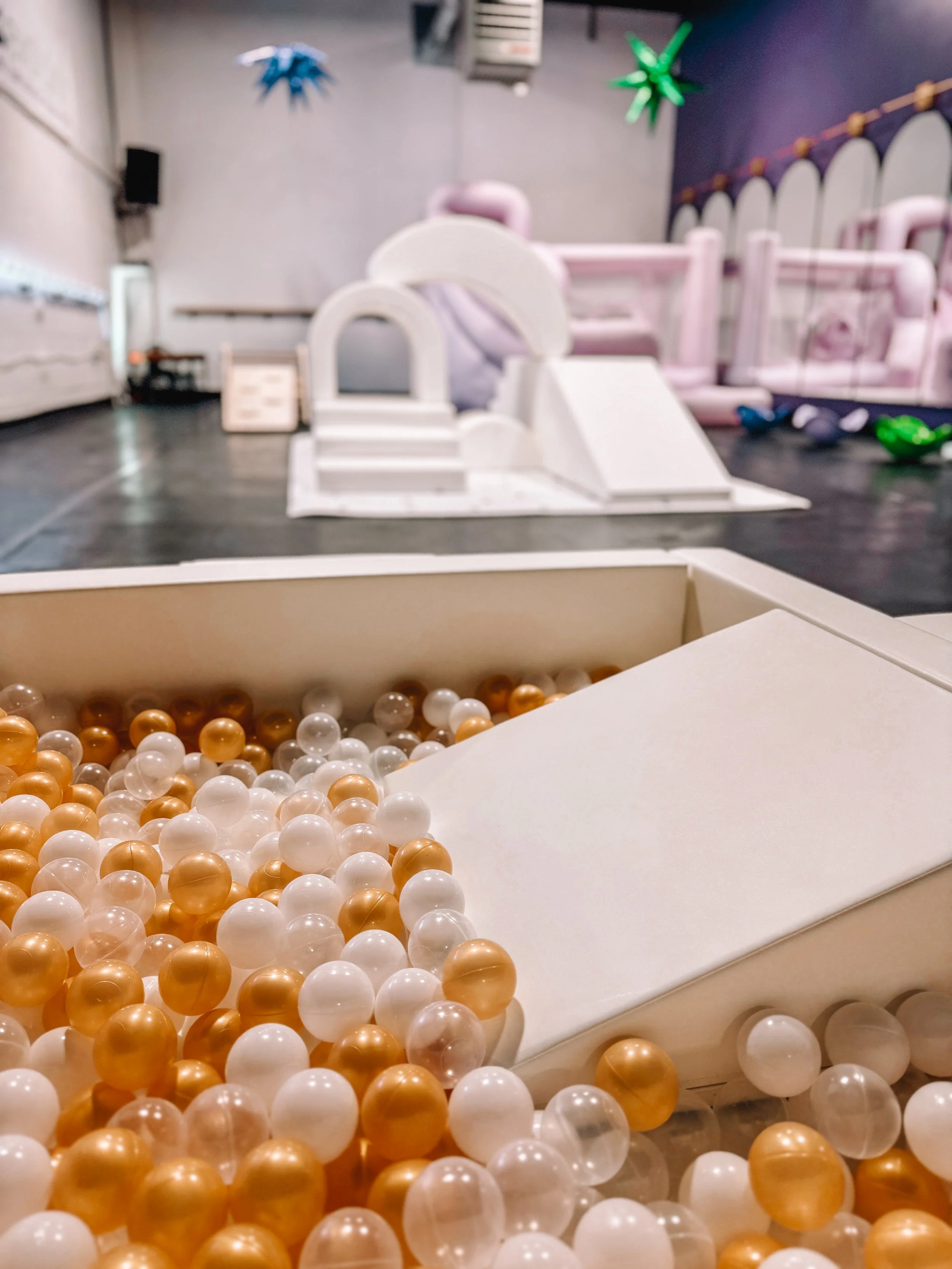 Indoor play area featuring a ball pit with white and gold plastic balls in the foreground and soft play structures, slides, and colorful hanging decorations in the background.