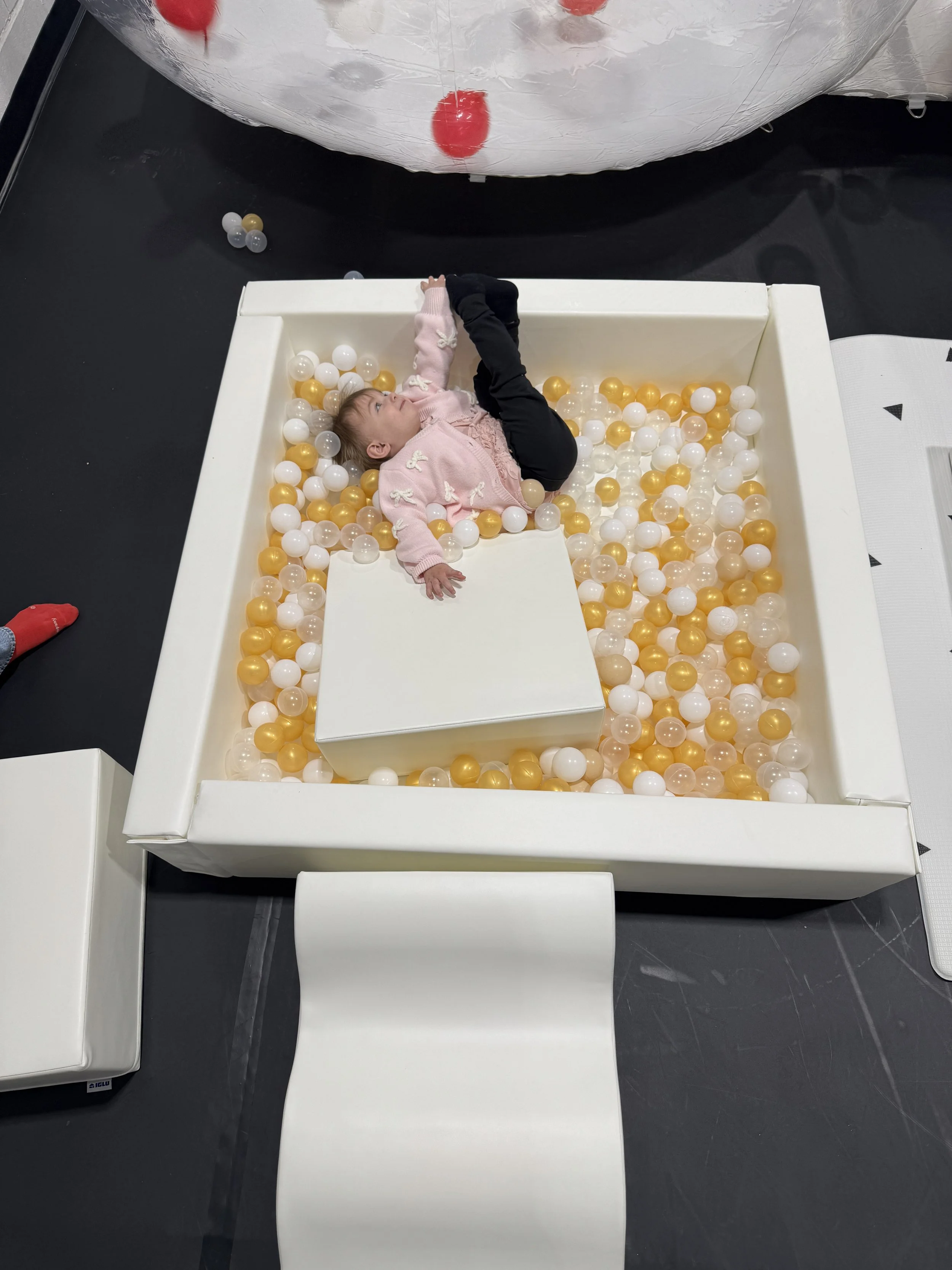 A young girl lying in a ball pit filled with white, gold, and transparent plastic balls. She is wearing a pink sweater with bows and black pants, and is looking up at the ceiling.