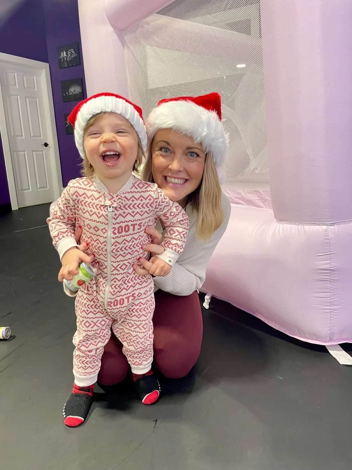 A woman and a young child wearing festive Santa hats, smiling and posing together indoors during a holiday celebration, with a pink inflatable structure in the background.