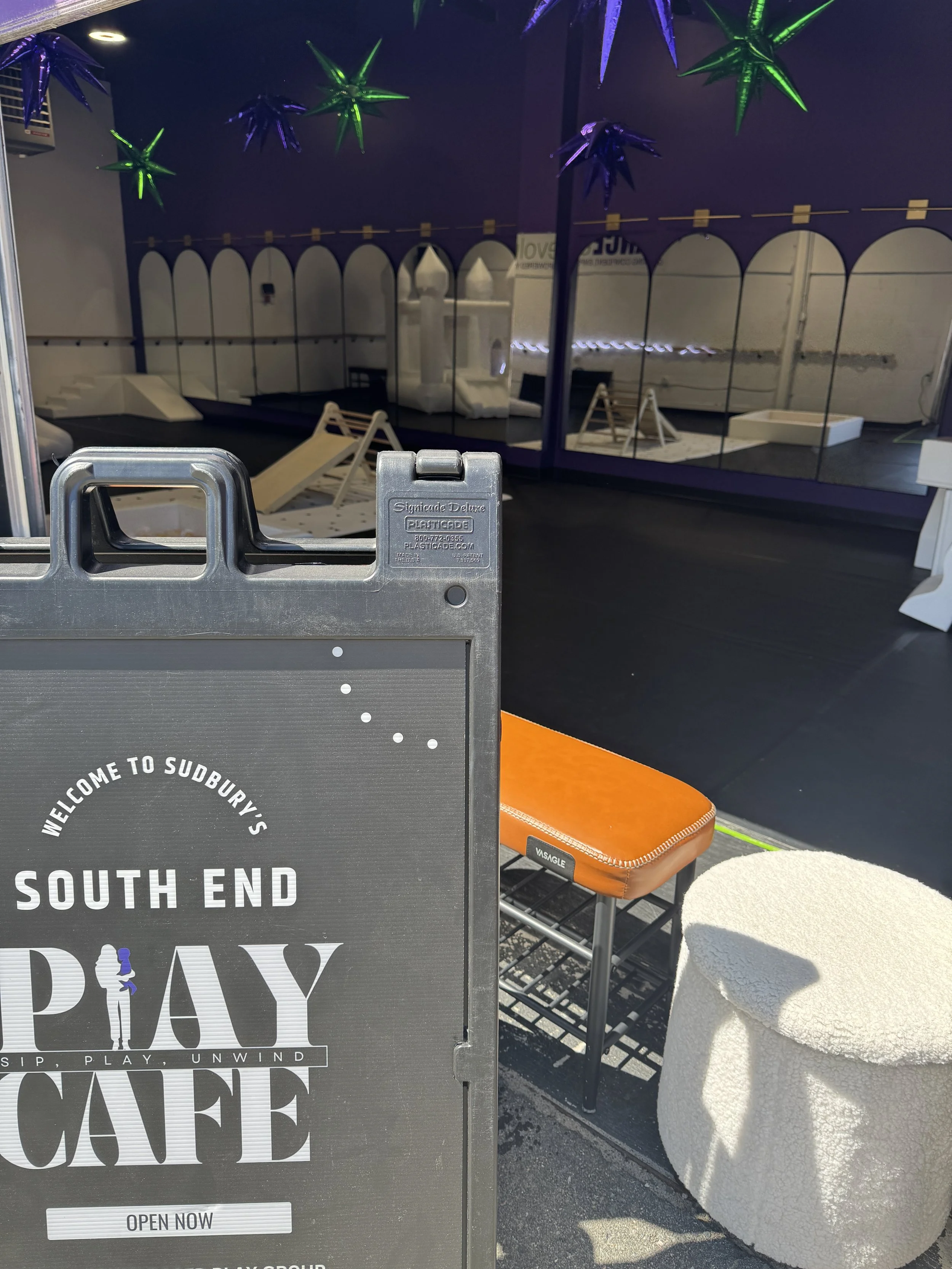 Signboard greeting visitors to South End Play Cafe with the phrase "Sip, Play, Unwind" and an illustration of a person holding a drink, inside a cafe with lounge chairs, a mirror wall, and decorative hanging stars.