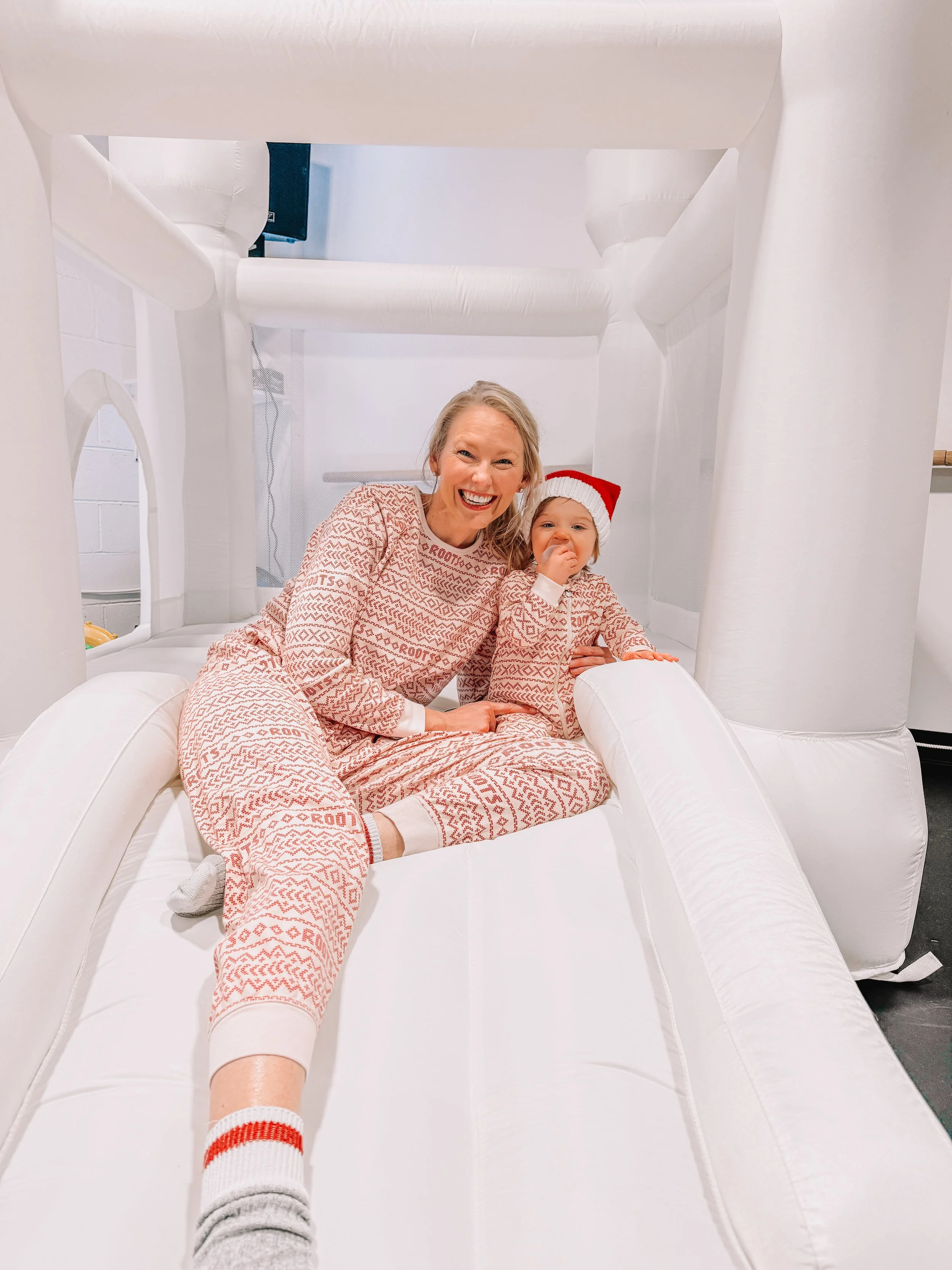 A woman and a young girl, both wearing matching holiday pajamas, playing on an inflatable slide. The girl is wearing a Santa hat and has her hand near her mouth. The woman is smiling and sitting nearby.