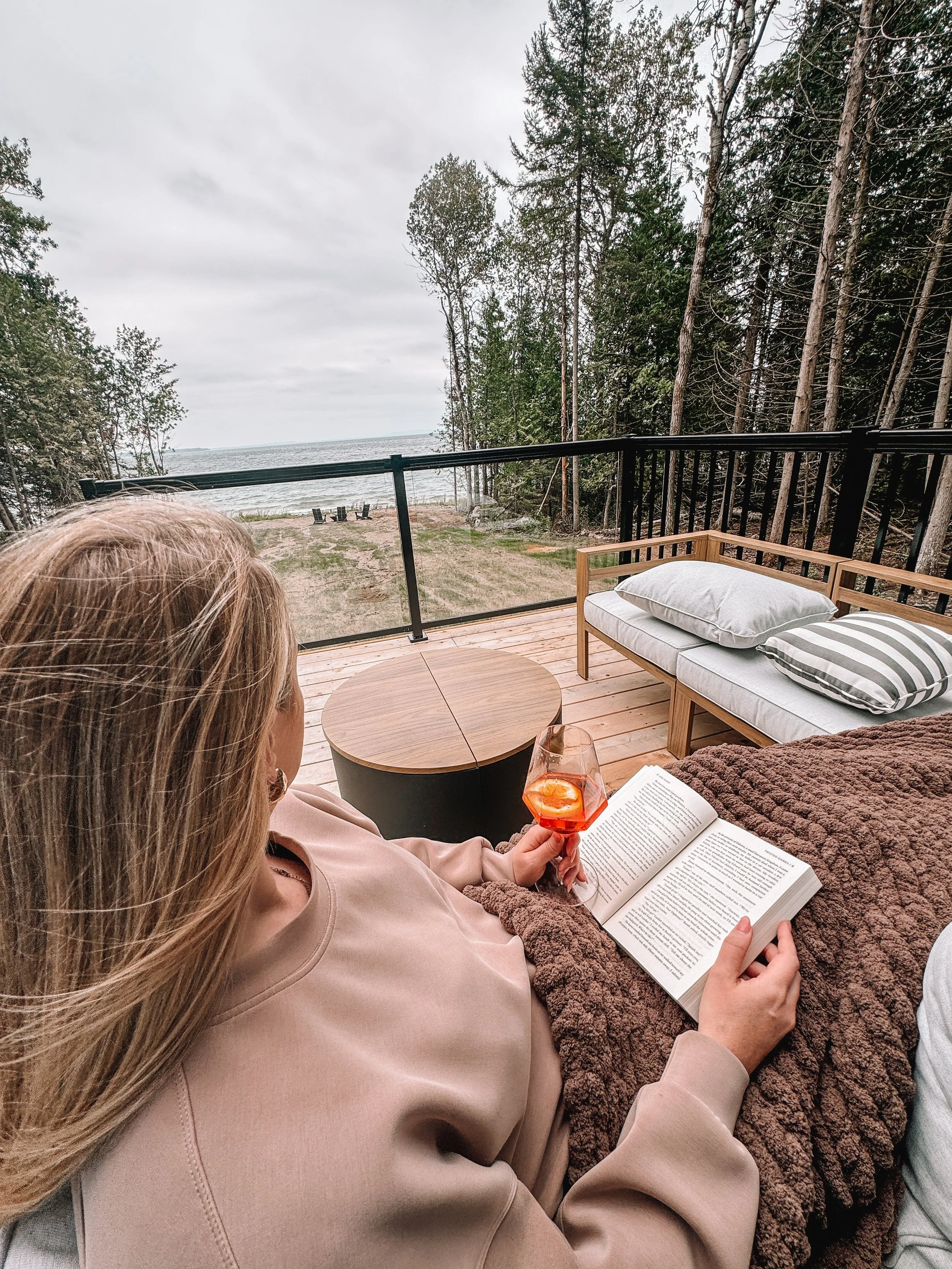 A woman relaxing on a porch sofa, reading a book and holding a glass of an orange-colored drink, with a view of a sandy beach, trees, and the ocean in the background.
