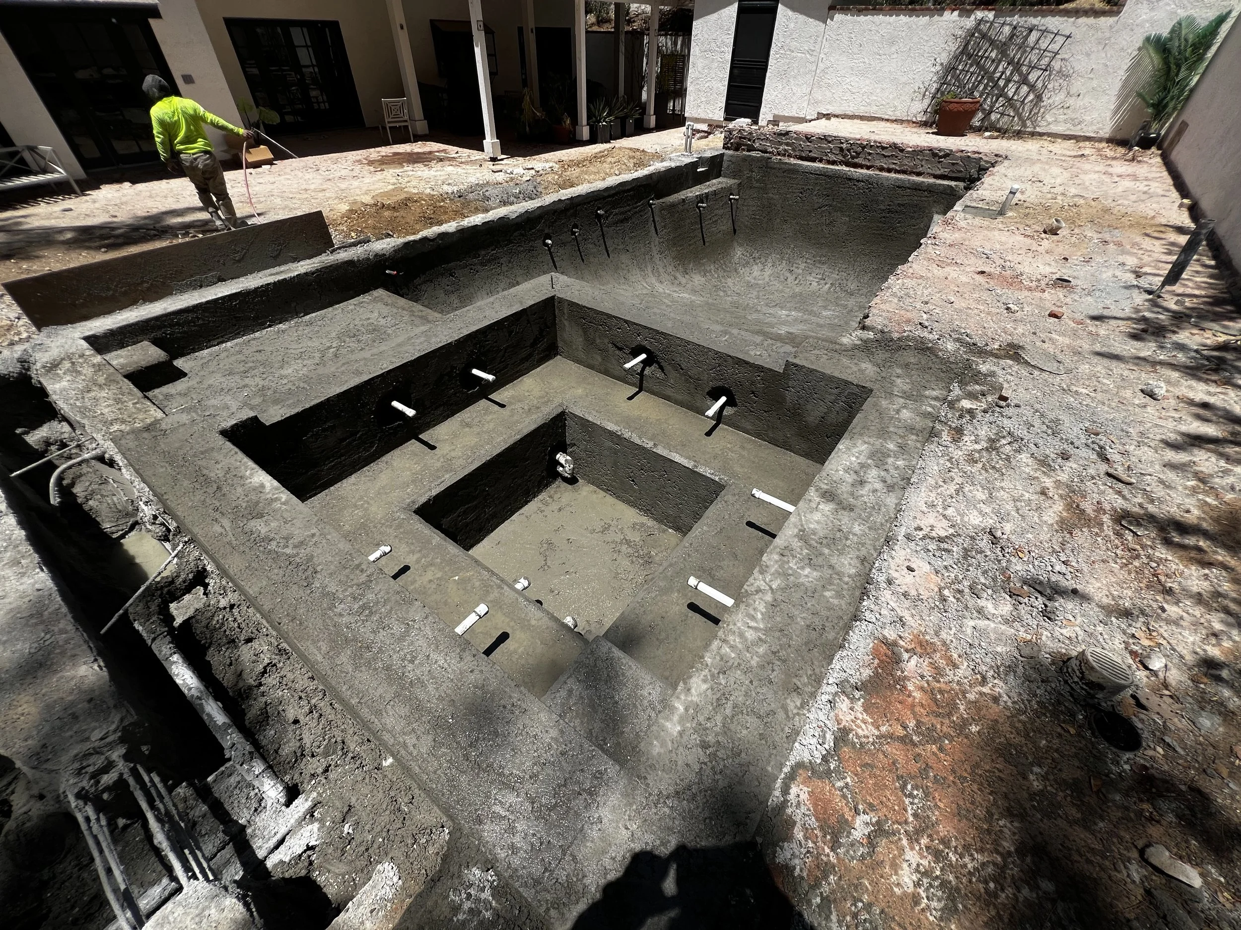 A construction worker in a high-visibility jacket working on a swimming pool excavation in a backyard. The pool outline is visible with rebar and plumbing fixtures inside.