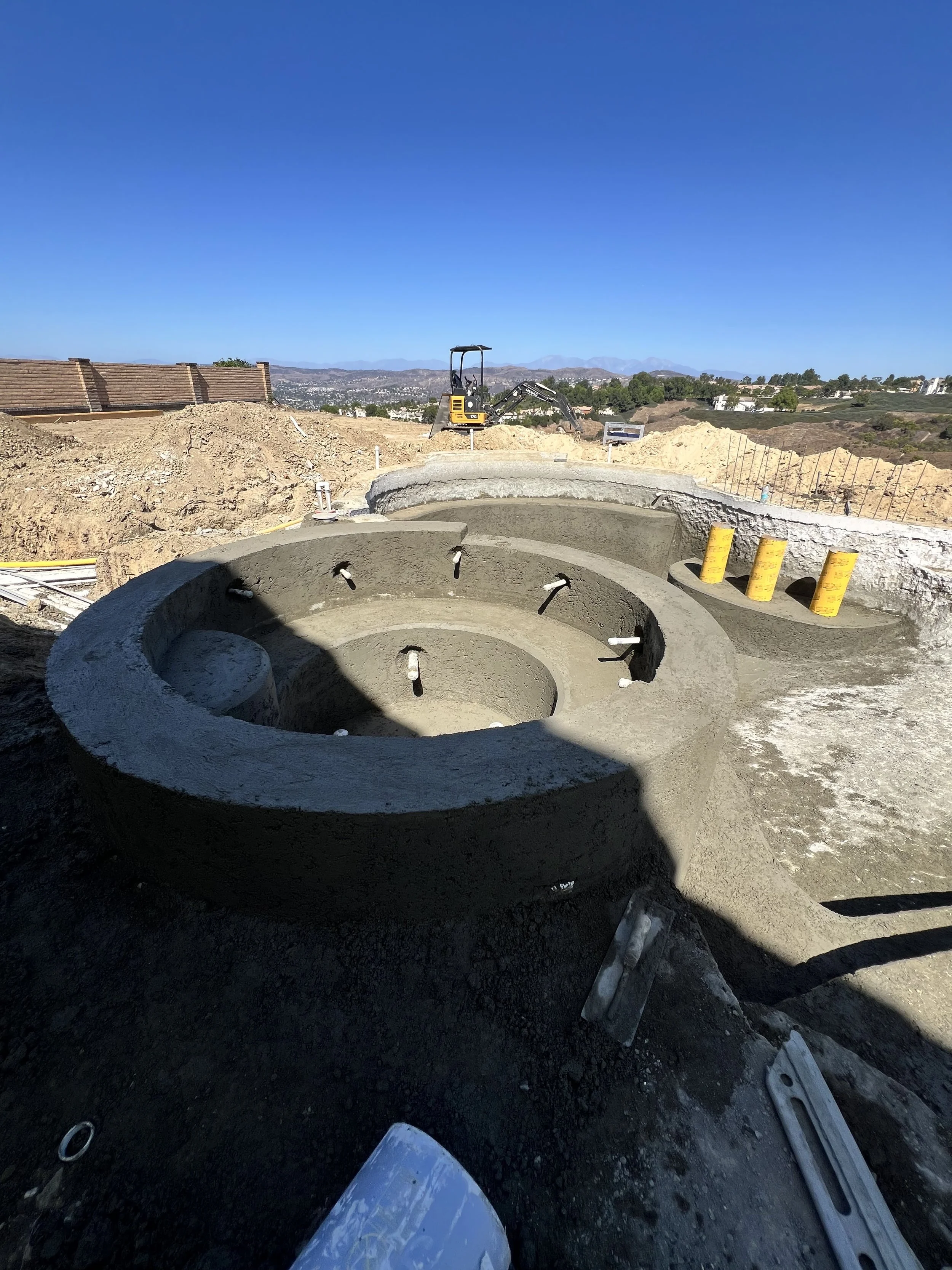 Construction site with circular foundation and excavator, in a hilly area with clear blue sky.