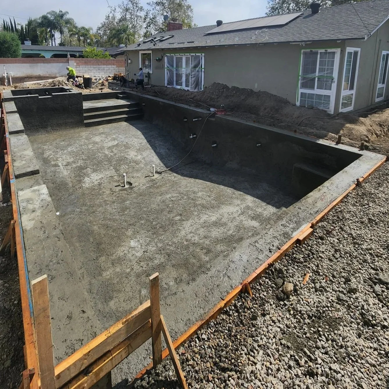Construction site of an in-ground swimming pool in a backyard, with a concrete pool shell built and workers working nearby, surrounded by a wooden fence and a house with solar panels on the roof.