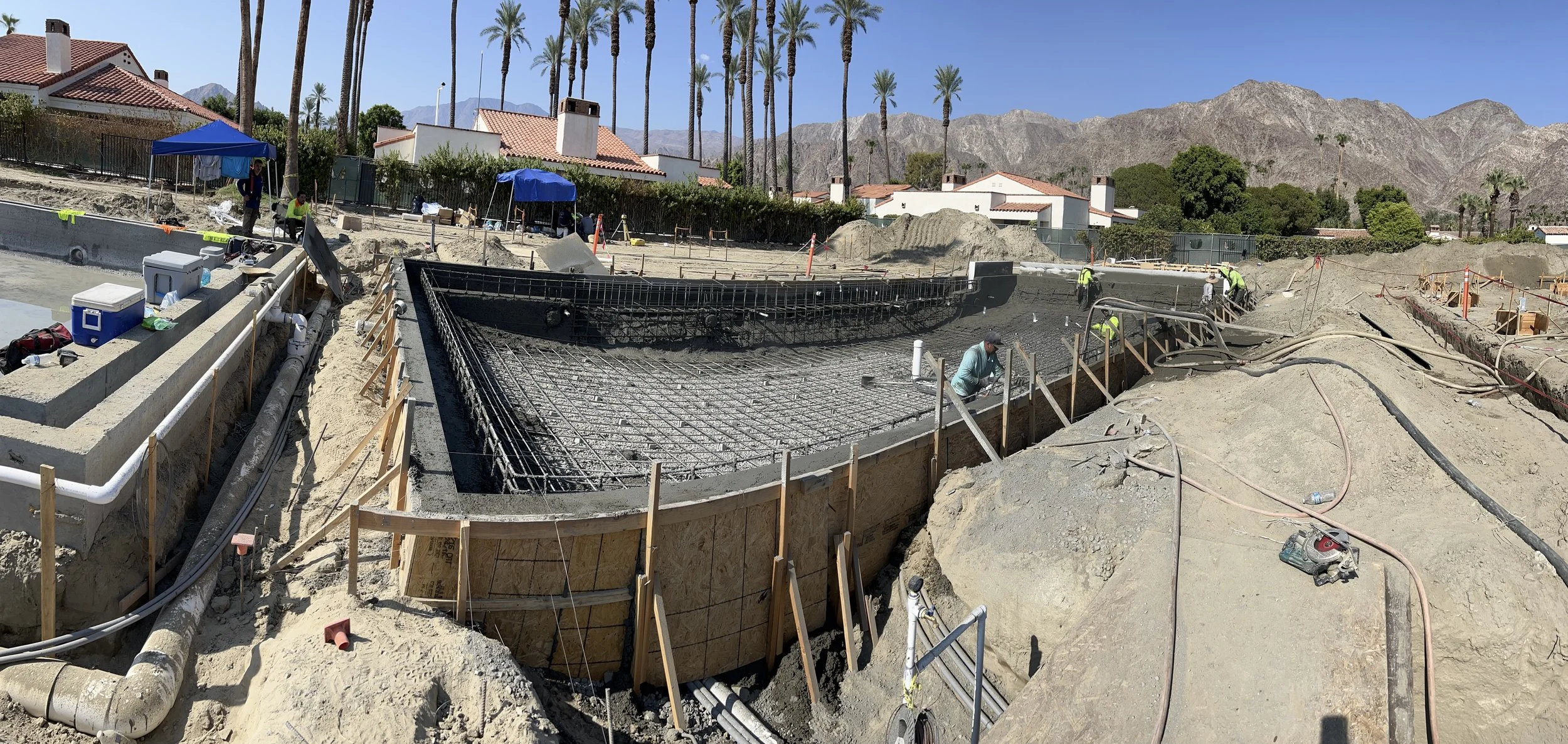 Construction workers building the foundation of a swimming pool in a residential backyard, with mountains and palm trees in the background.