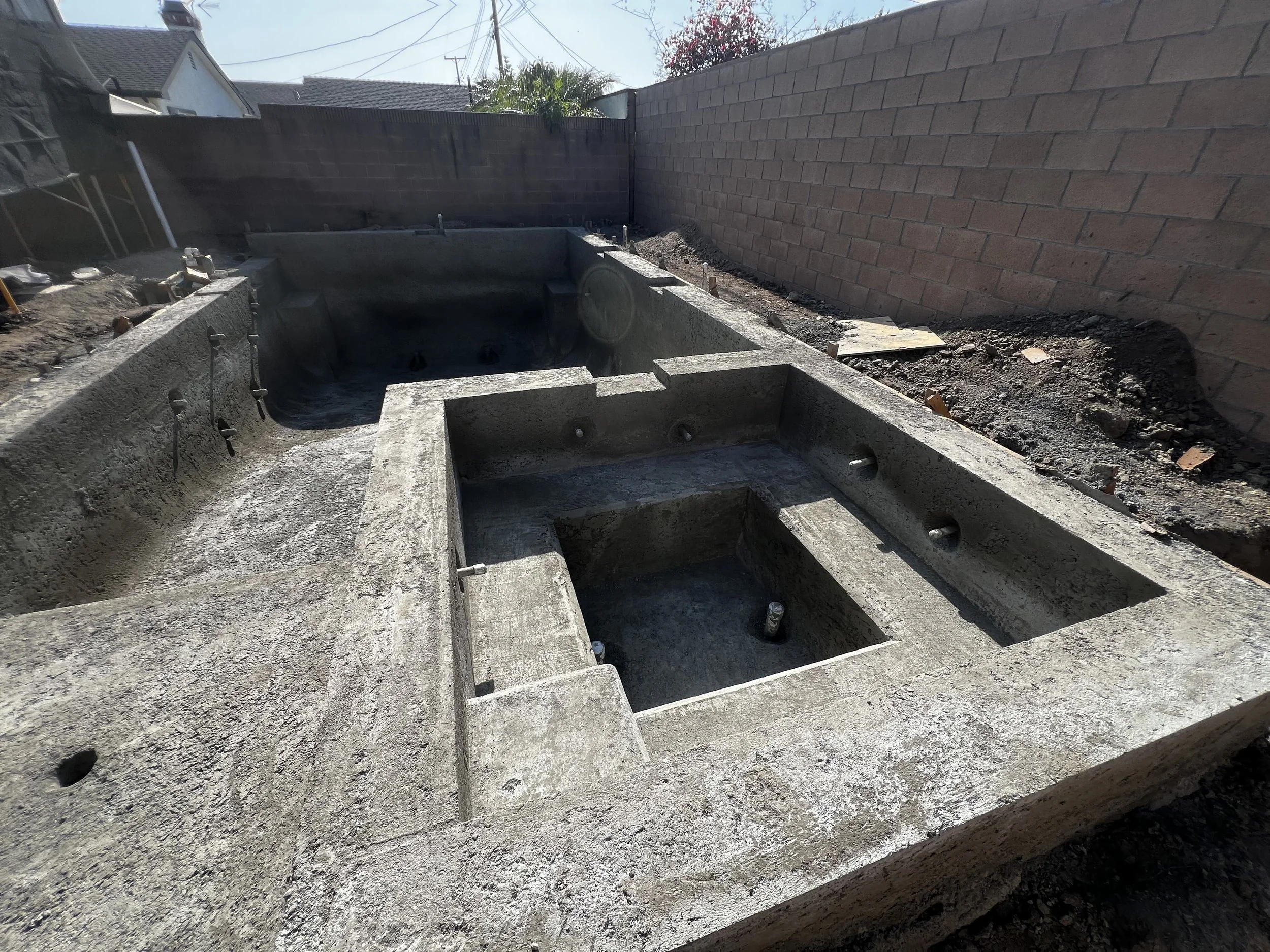 Under construction backyard with concrete spa, brick wall, and surrounding dirt.