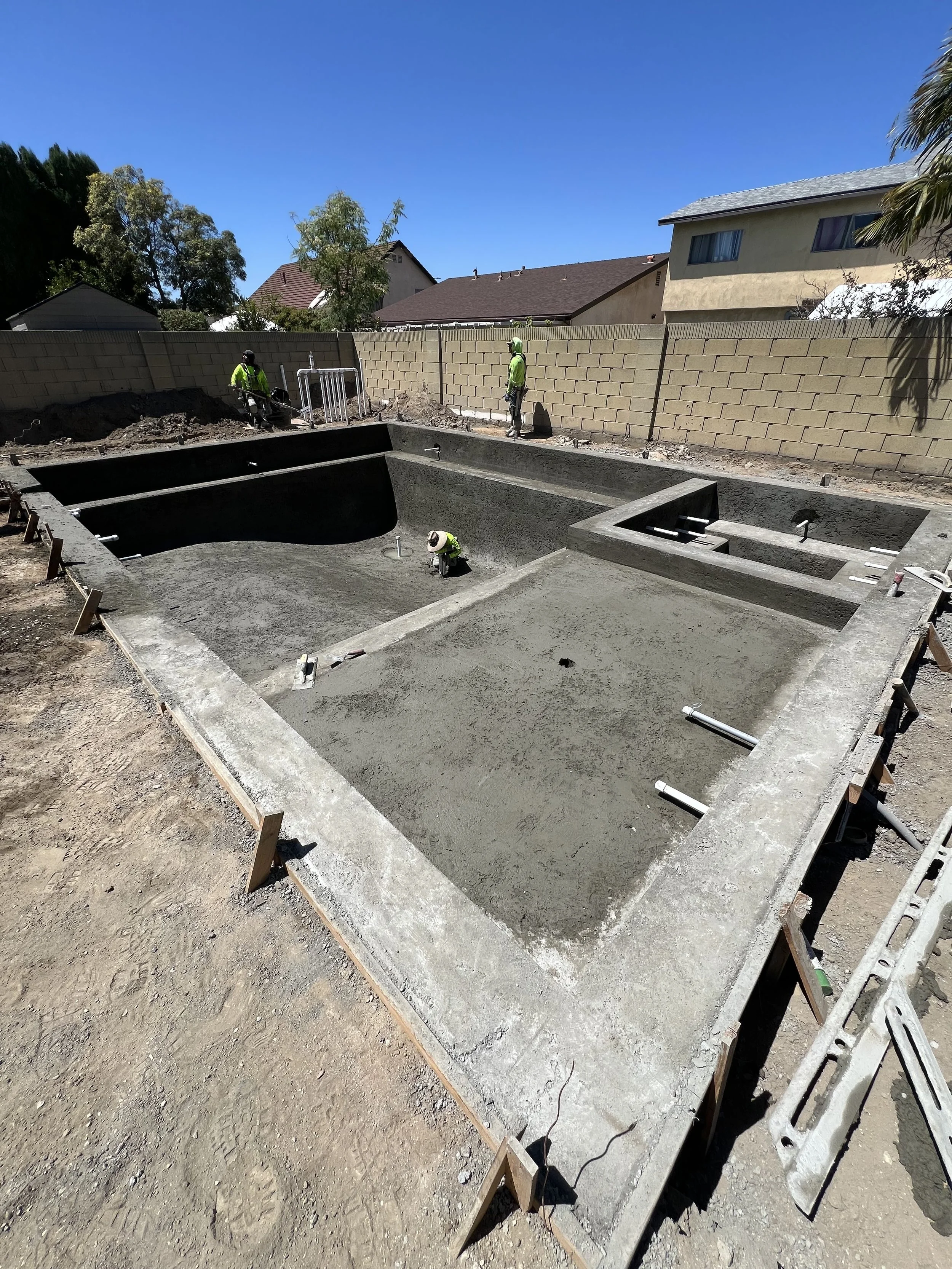 Construction site with concrete pool and workers wearing yellow safety vests and helmets, in a residential backyard with a brick wall and houses in the background.