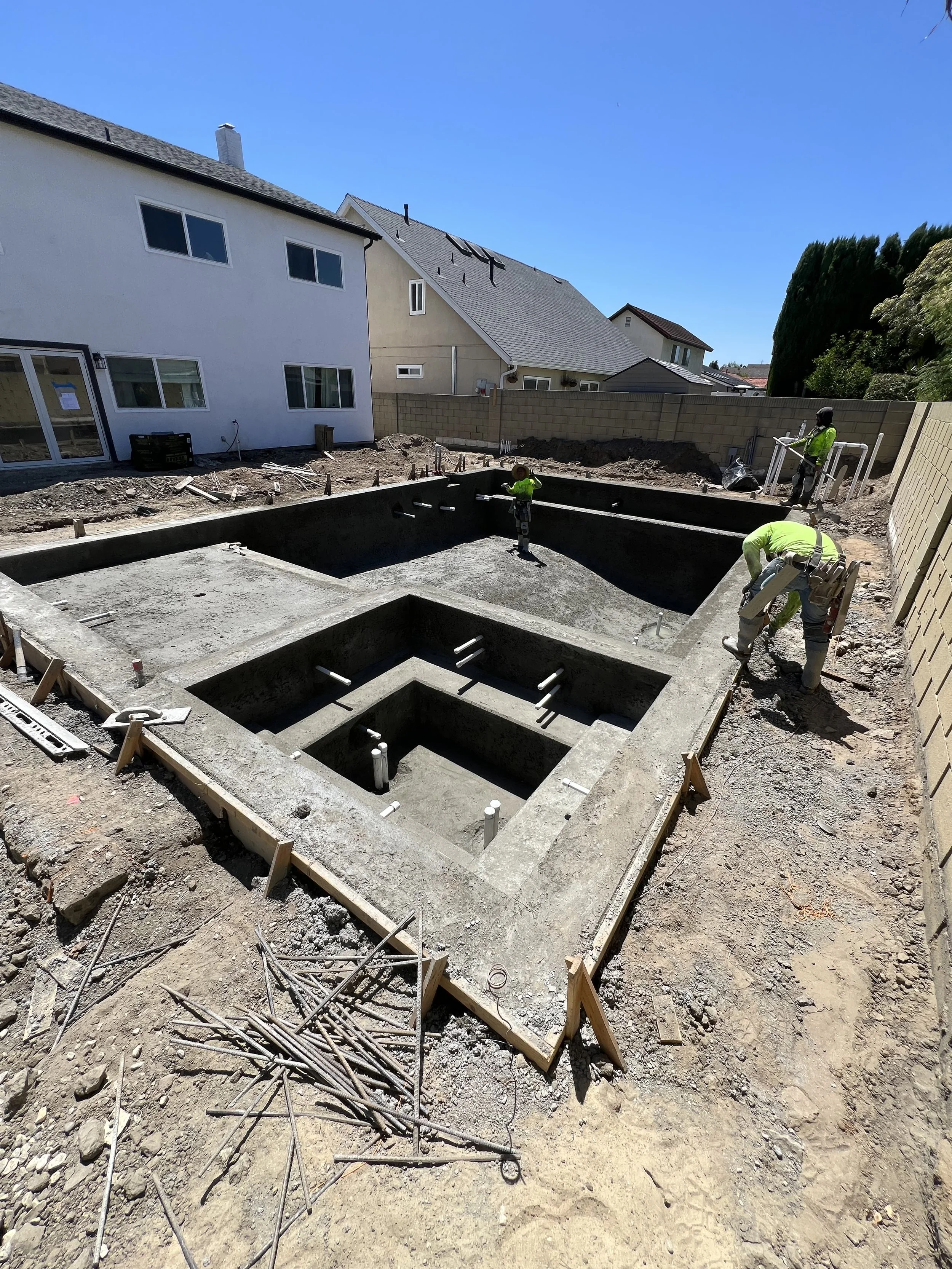 Construction workers are building a pool in a backyard, with concrete pool shells and construction materials around.