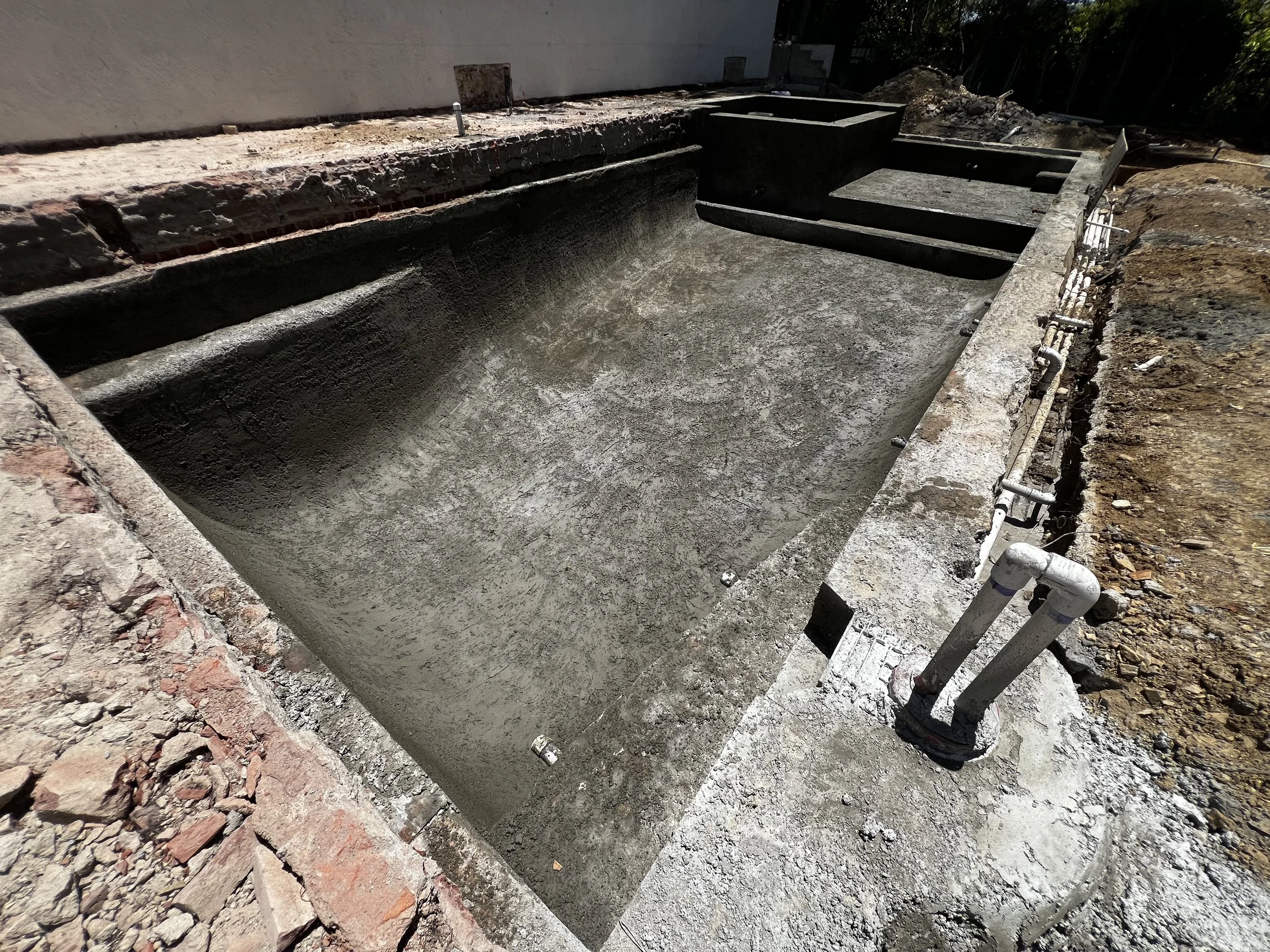 Construction site with a concrete pool or pond under development, surrounded by exposed soil, bricks, and construction pipes.