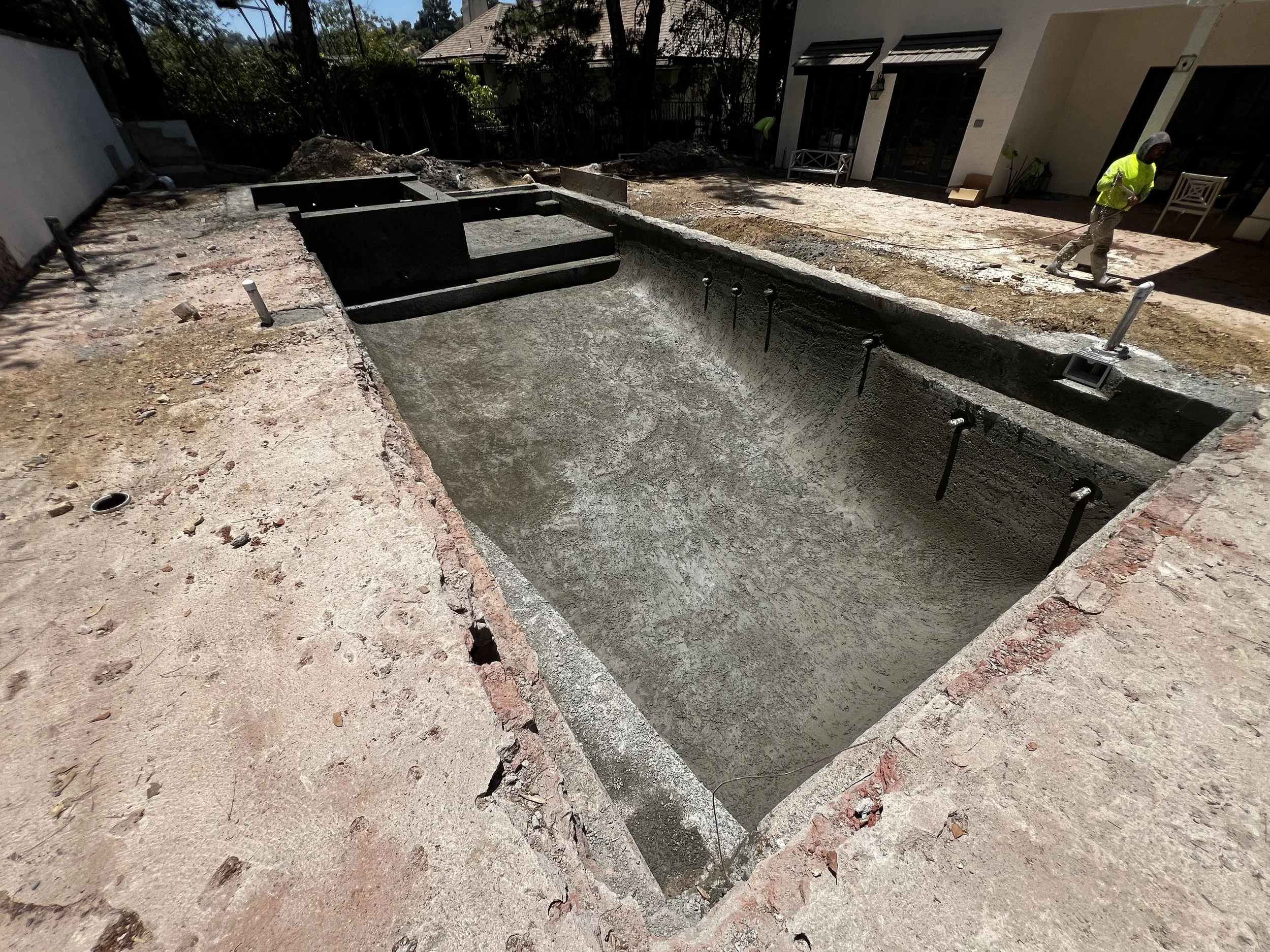 A backyard with an empty, in-ground swimming pool under construction, surrounded by a partially finished patio. A worker wearing a neon yellow safety vest and white hat is standing on the patio, which is being prepared for paving.