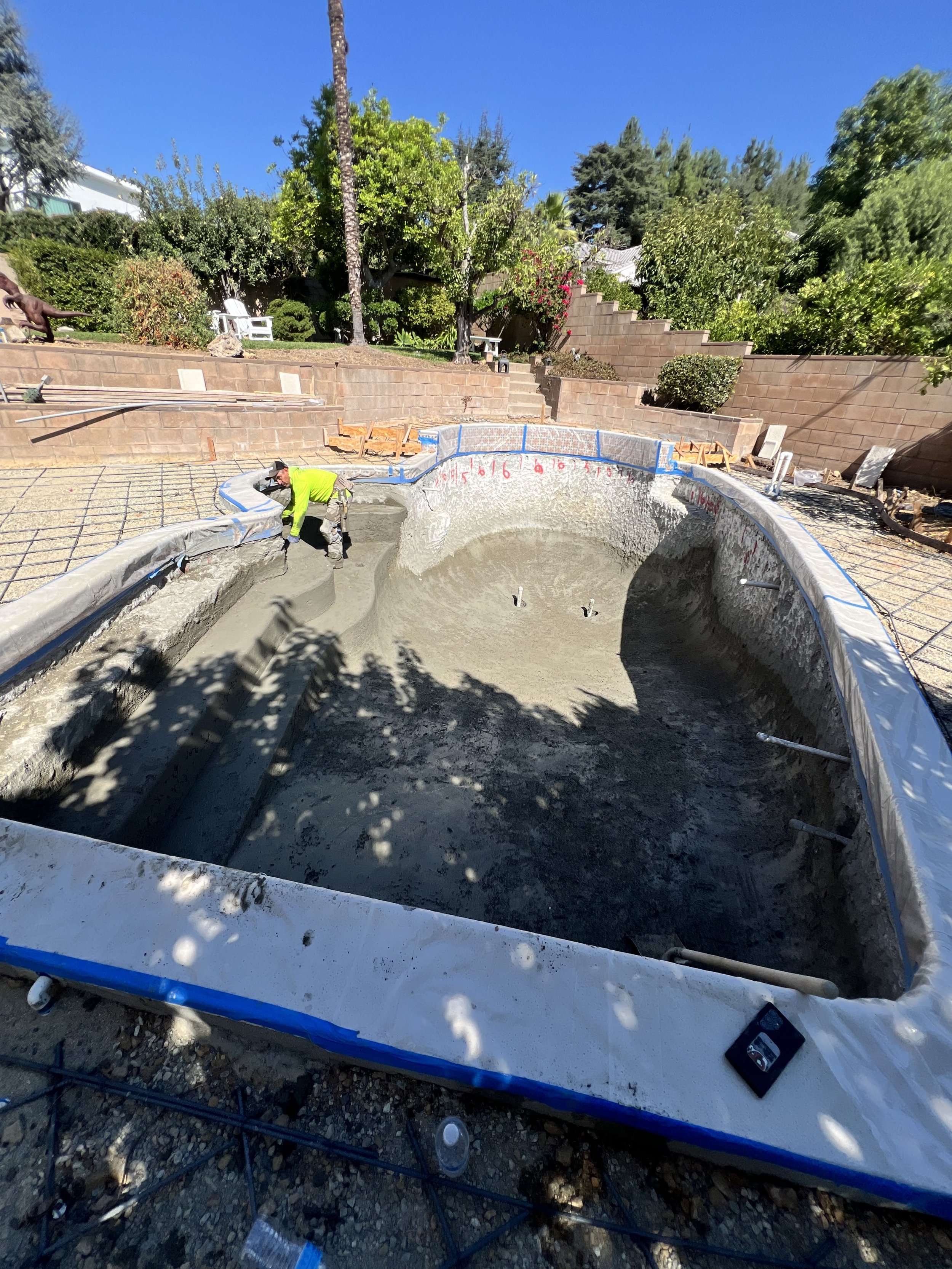Construction worker smoothing cement in a pool under a clear blue sky, with trees and a brick wall in the background.