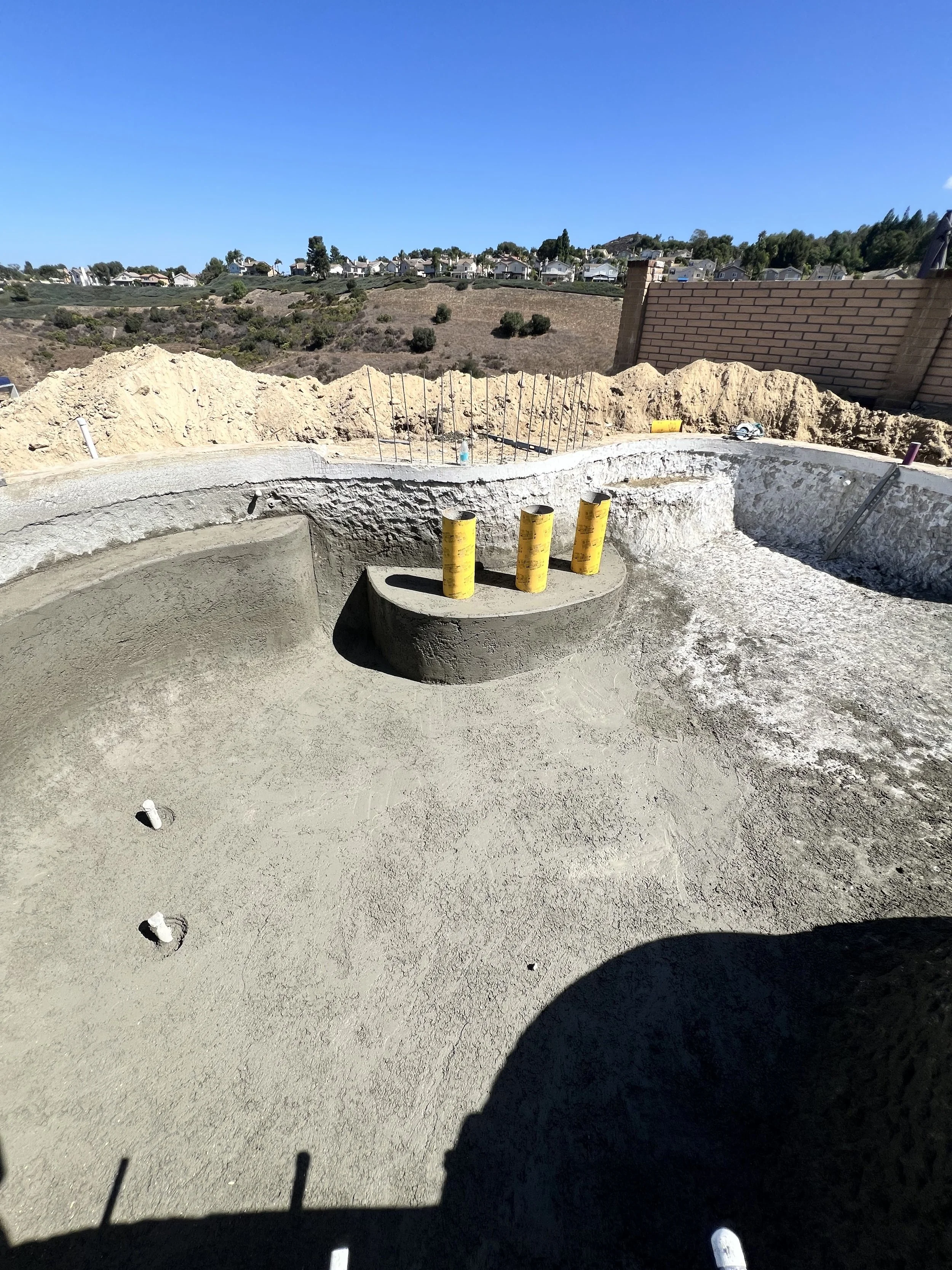 Construction site showing a curved concrete foundation with three yellow pipes sticking out, surrounded by dirt and construction debris, with a brick wall and hillside with houses in the background.