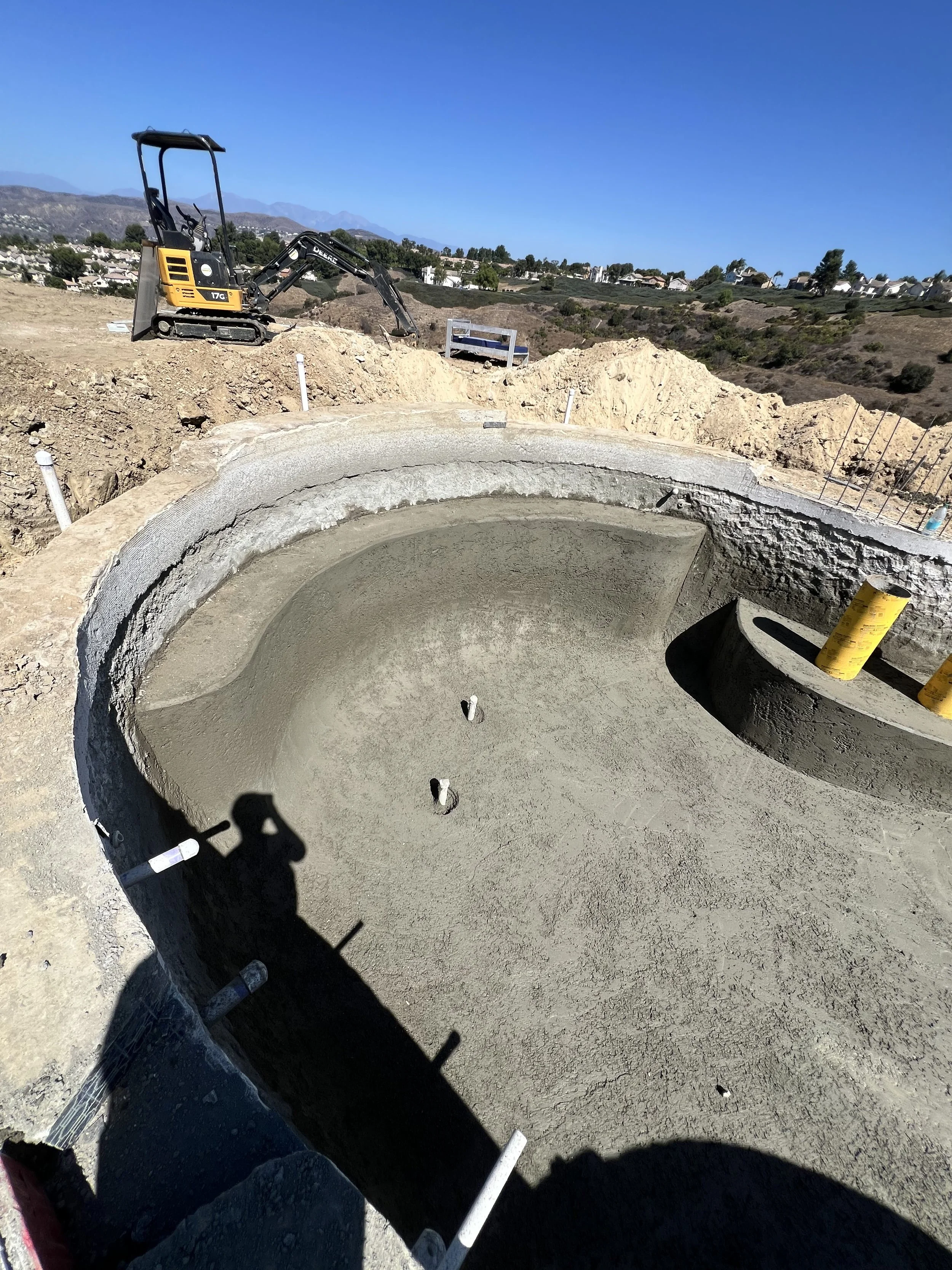 Construction site showing a large circular concrete foundation with a small excavator nearby, set in a hilly landscape with houses in the background and clear blue sky.