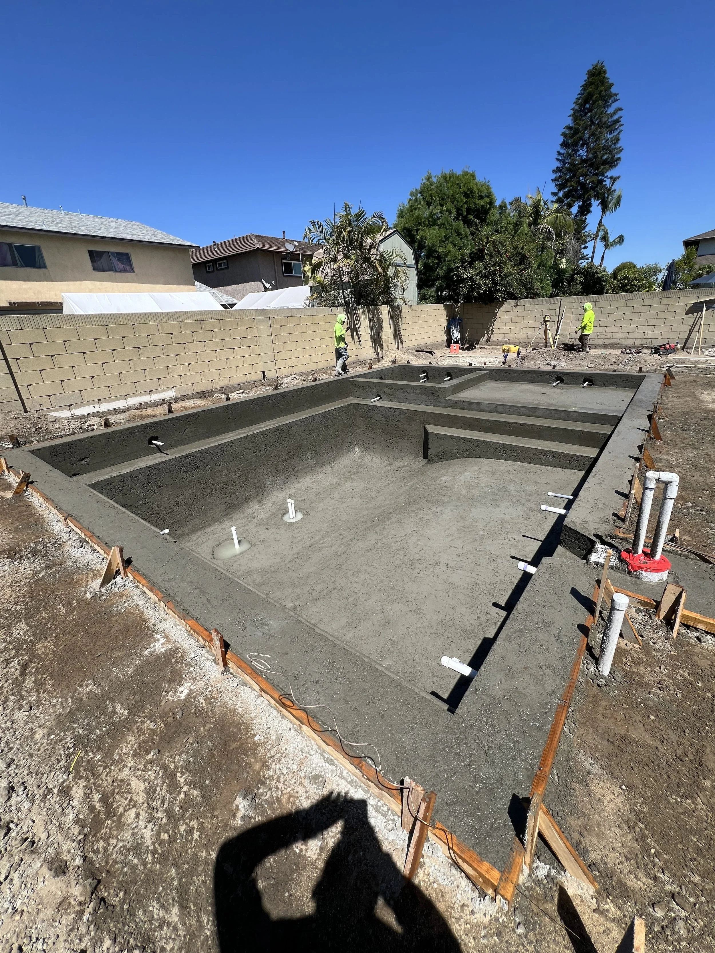 Construction site with a in-ground swimming pool being built, two workers in bright yellow shirts, a brick wall in the background, and a clear blue sky.