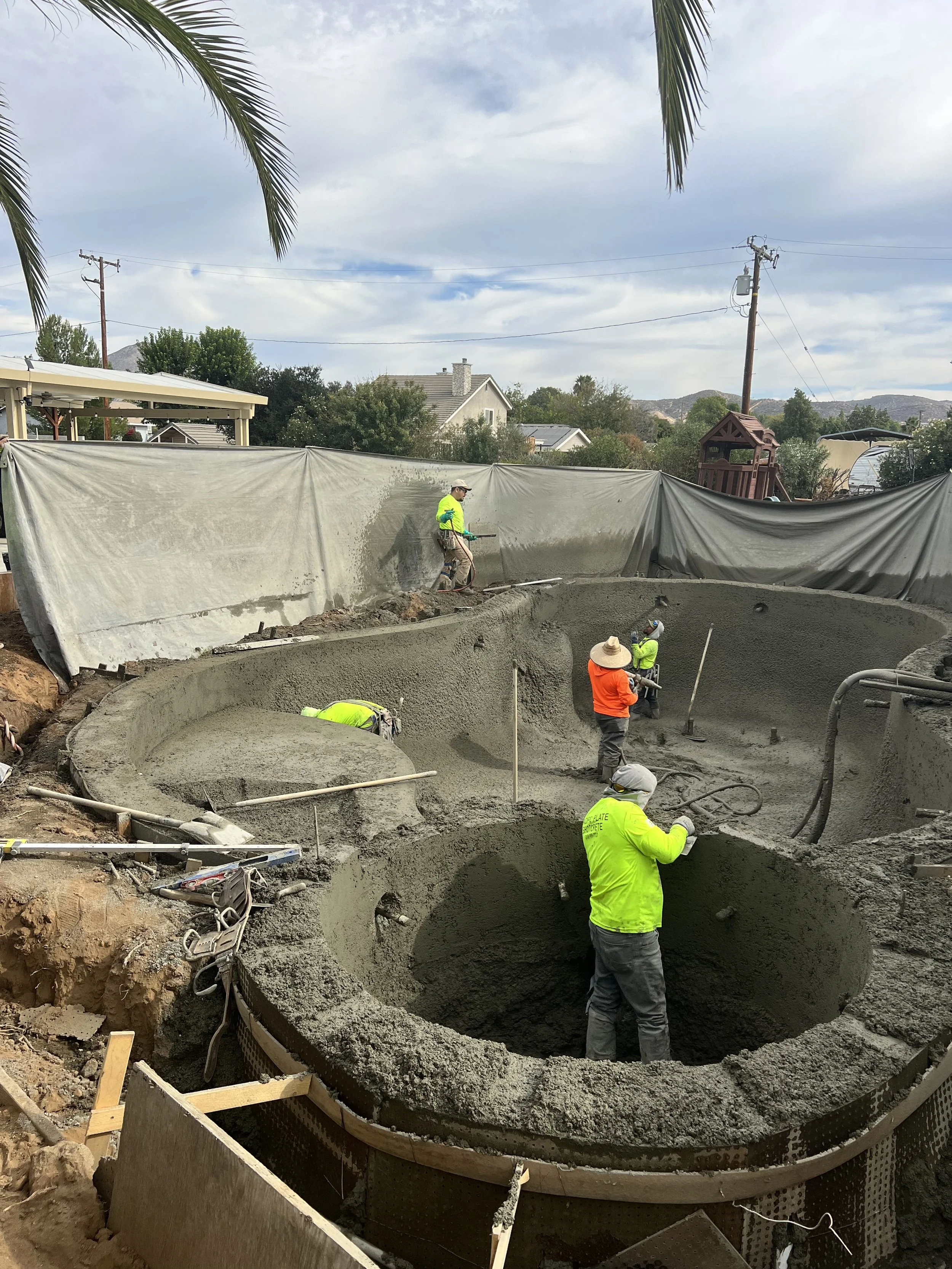 Construction workers pouring concrete into a round pool structure at a construction site, with a cloudy sky and residential houses in the background.
