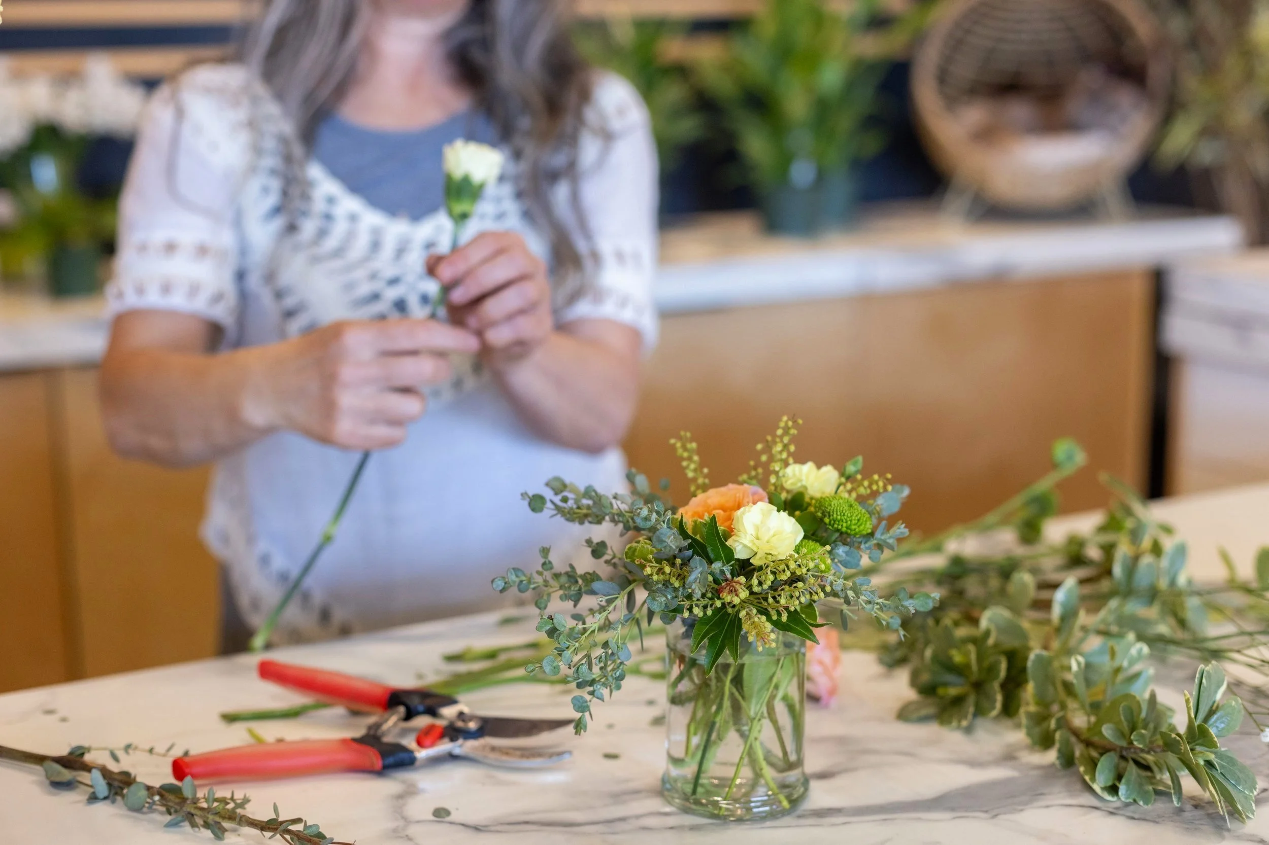 Woman arranging a floral bouquet with yellow and orange flowers, greenery, and eucalyptus on a marble table.