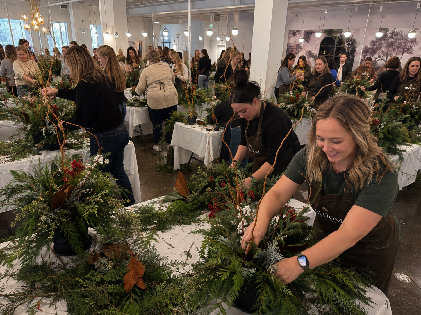 People gather in a bright, spacious room to create holiday floral arrangements with greenery, red berries, and decorative branches. Some are smiling and focused on their craft, wearing aprons.