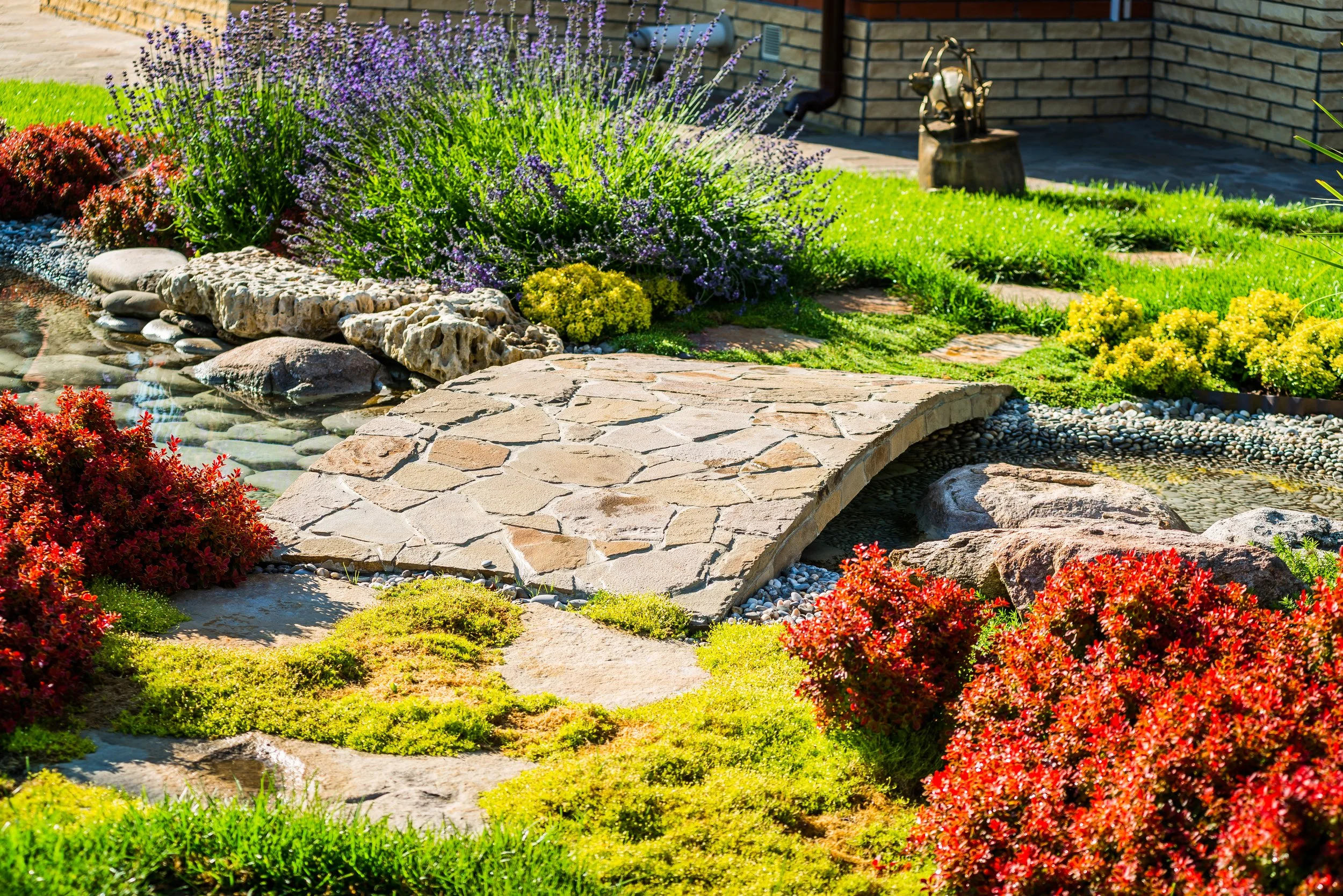 Colorful garden with stone pathway, purple flowering plants, red and yellow bushes, green grass, small pond with rocks, and a brick wall in the background.