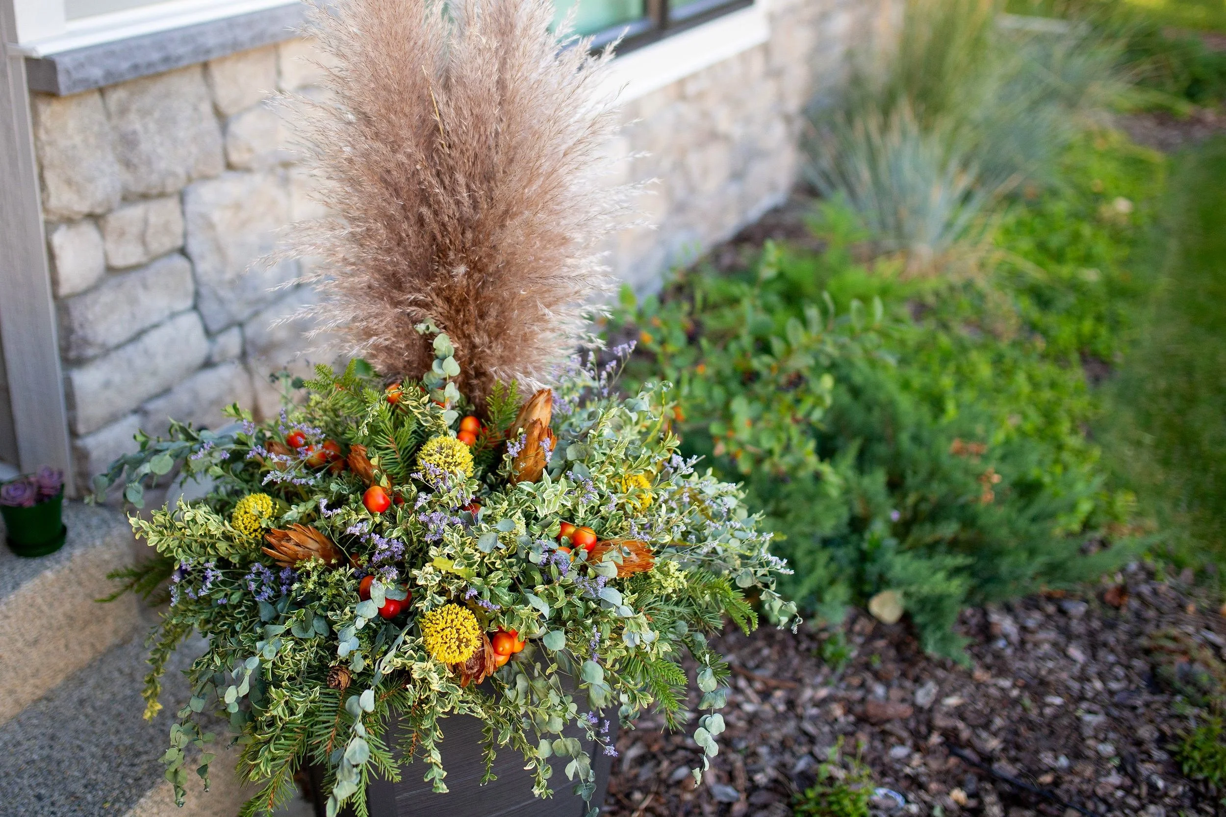 Decorative autumn planter with pampas grass, orange berries, yellow flowers, and green foliage outside near stone wall and garden bed.