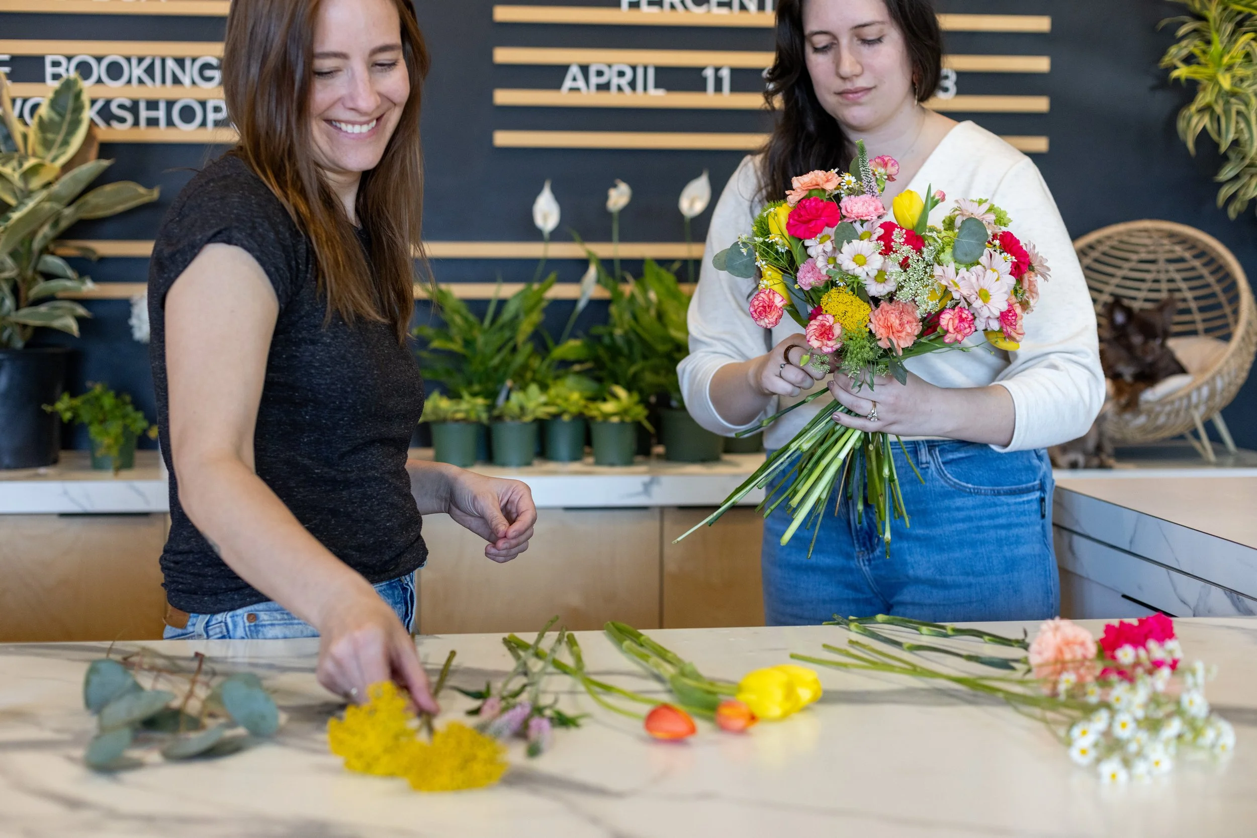 Two women arranging colorful flowers at a flower shop, with plants on shelves and a sign in the background.