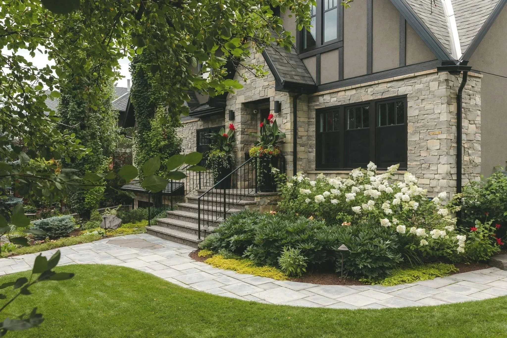A residential house with a stone facade, black window frames, and a staircase leading to the front entrance, surrounded by a lush, well-maintained garden with green shrubs, white and red flowers, and a paved pathway.
