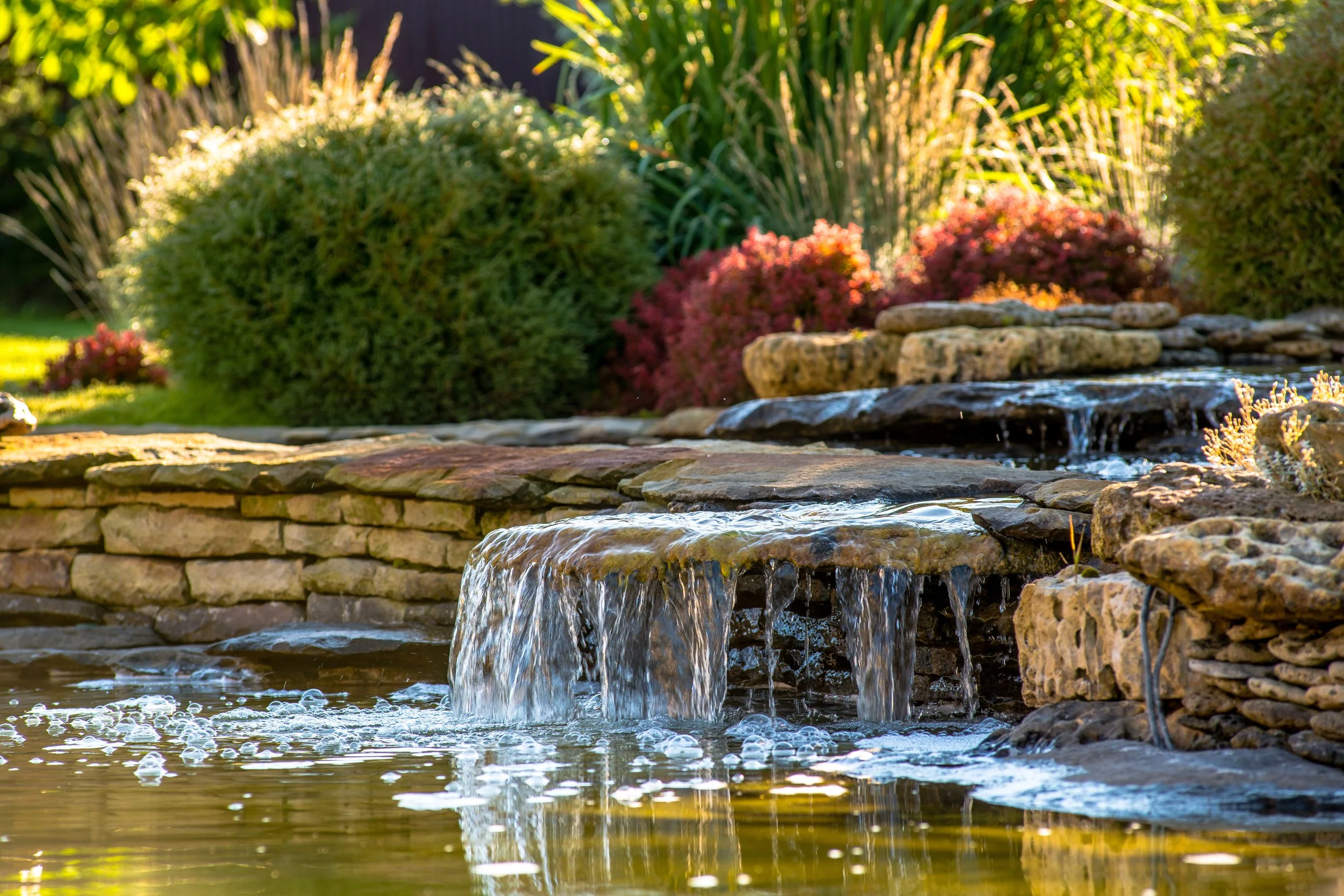 Waterfall cascading over stacked stones in a garden pond with lush greenery and colorful bushes in the background.