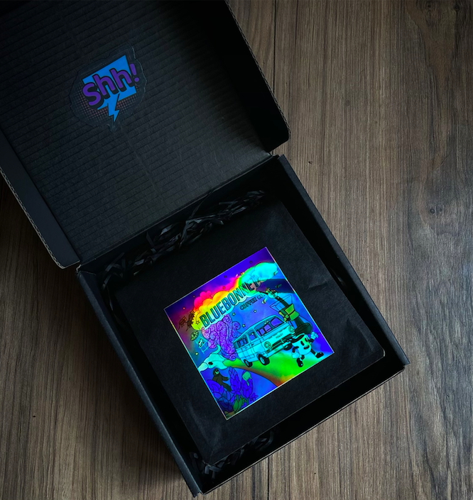 Open black box with a colorful sticker or card inside, featuring a psychedelic design including a van and text. The box's lid has a "shh!" logo with a lightning bolt.