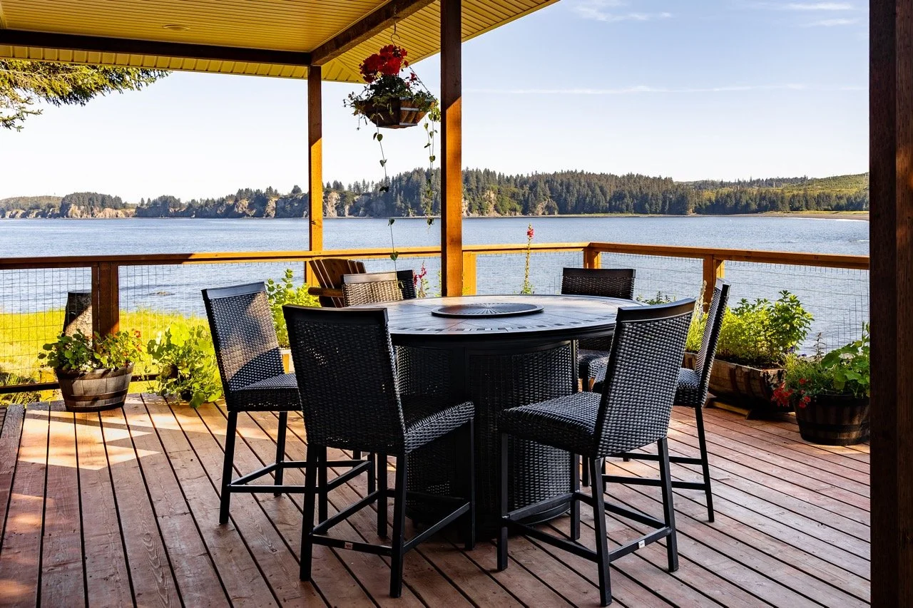 A deck overlooking a ocean with a round black outdoor table and six matching chairs, potted plants, and hanging flower baskets, with hills and trees in the distance under a partly cloudy sky.