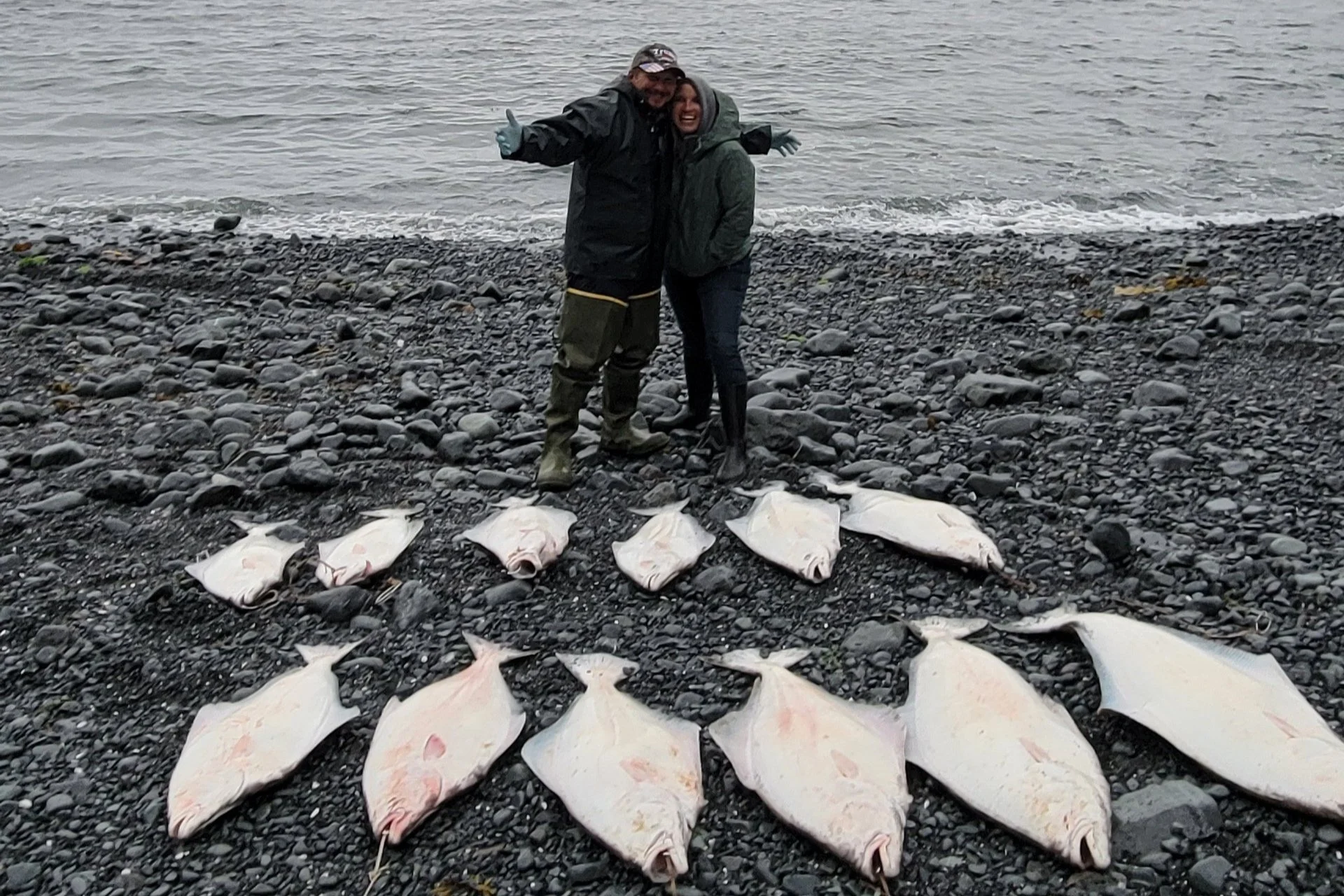 Two people standing on a rocky shoreline, surrounded by fishing nets and fish, with the ocean in the background.