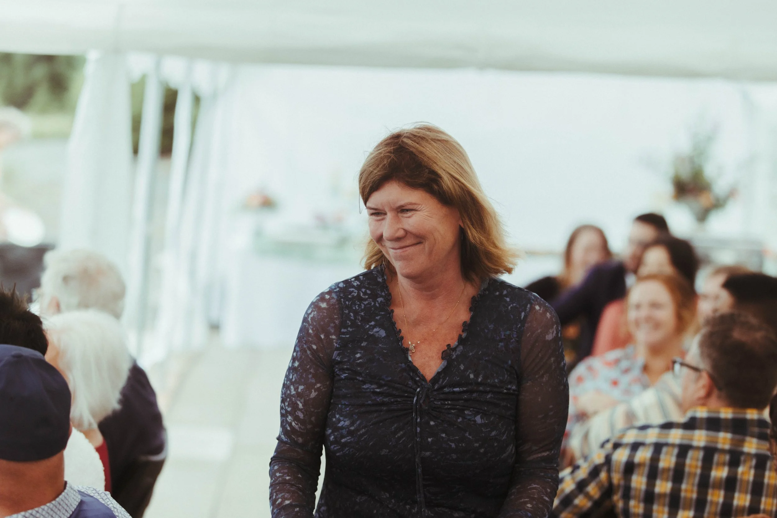 A woman with shoulder-length reddish-brown hair smiling and looking slightly to the side at an indoor social gathering.