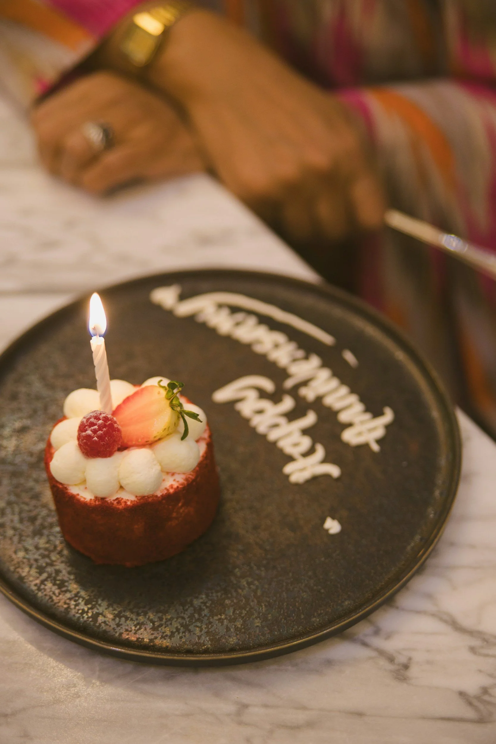 Close-up of a small birthday cake with a lit candle, strawberries, and cream on top, placed on a black plate with white lettering, with a person in colorful clothing in the background.