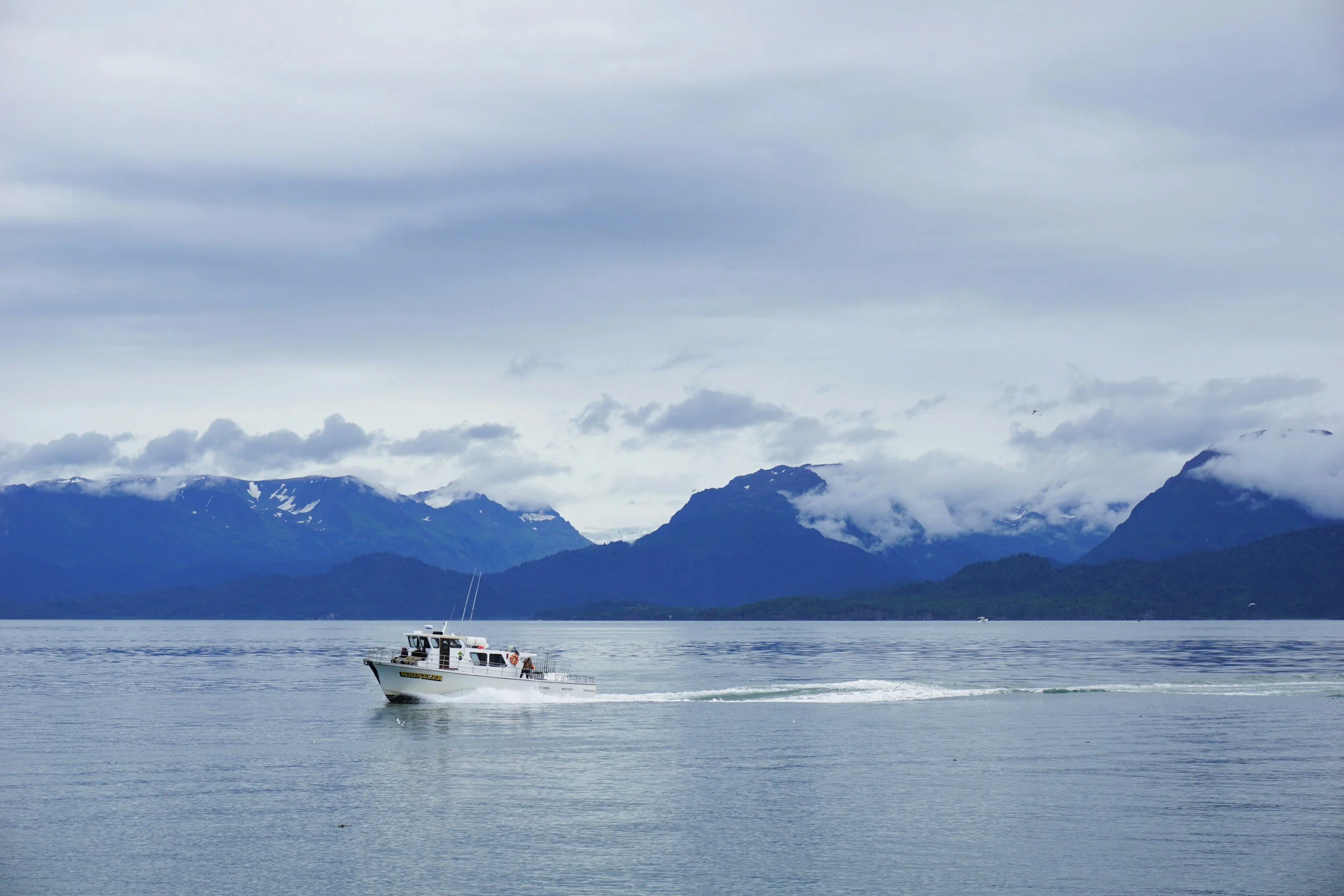 A white boat traveling across a calm body of water with mountain ranges and cloudy sky in the background.