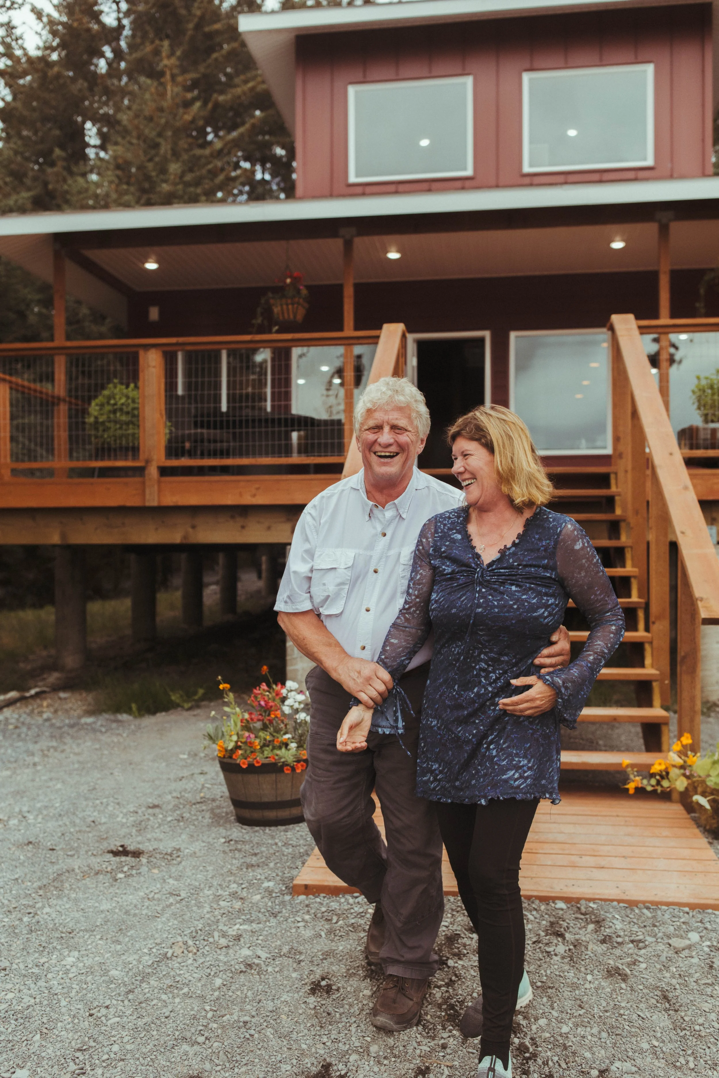 A man and woman standing together outside a two-story house with a red exterior and large windows, in front of a wooden deck with stairs, during daytime.