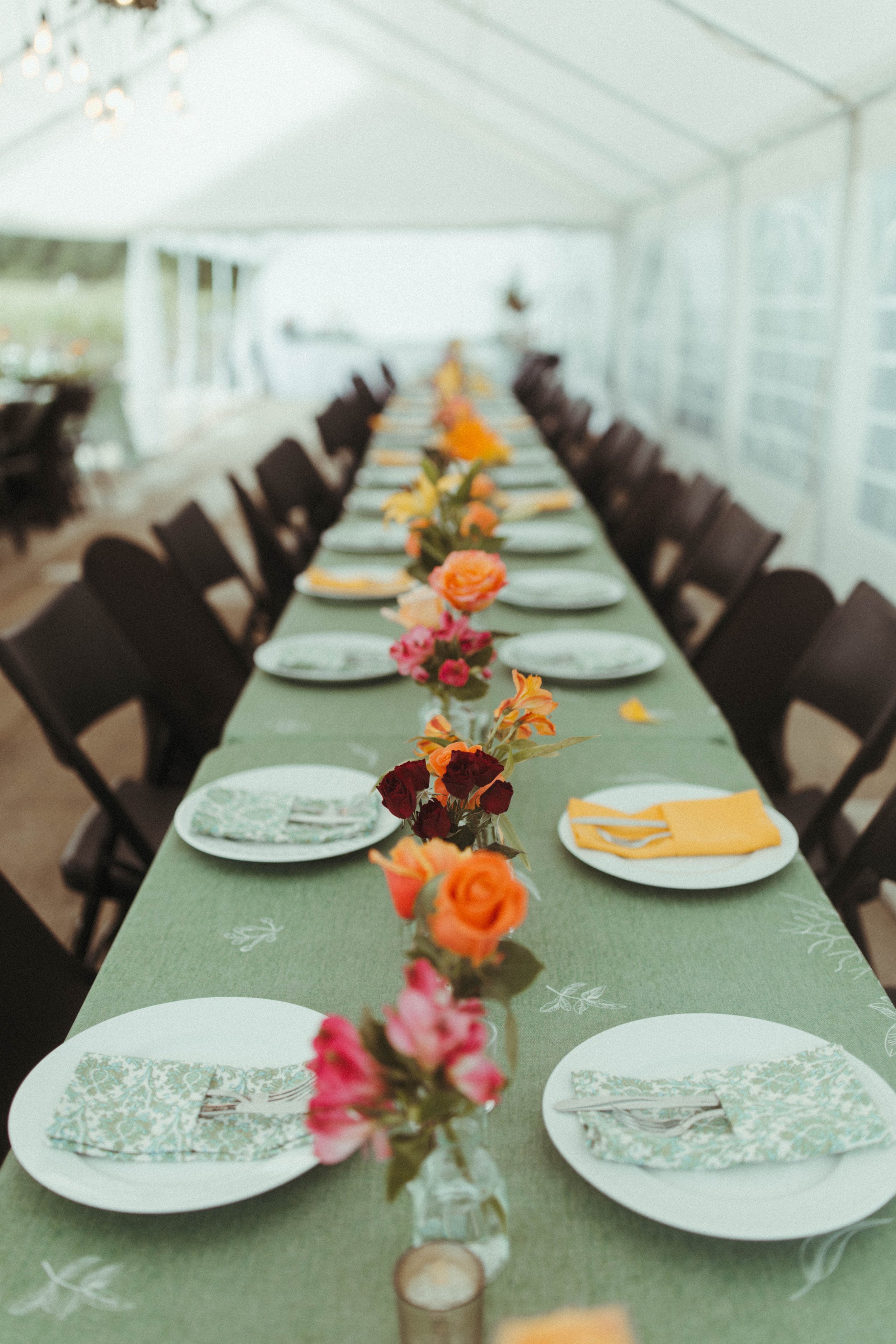 Long rectangular dining table with green tablecloth, decorated with a row of colorful flowers in small vases, set with white plates, patterned napkins, and silverware, under a white tent with string lights.